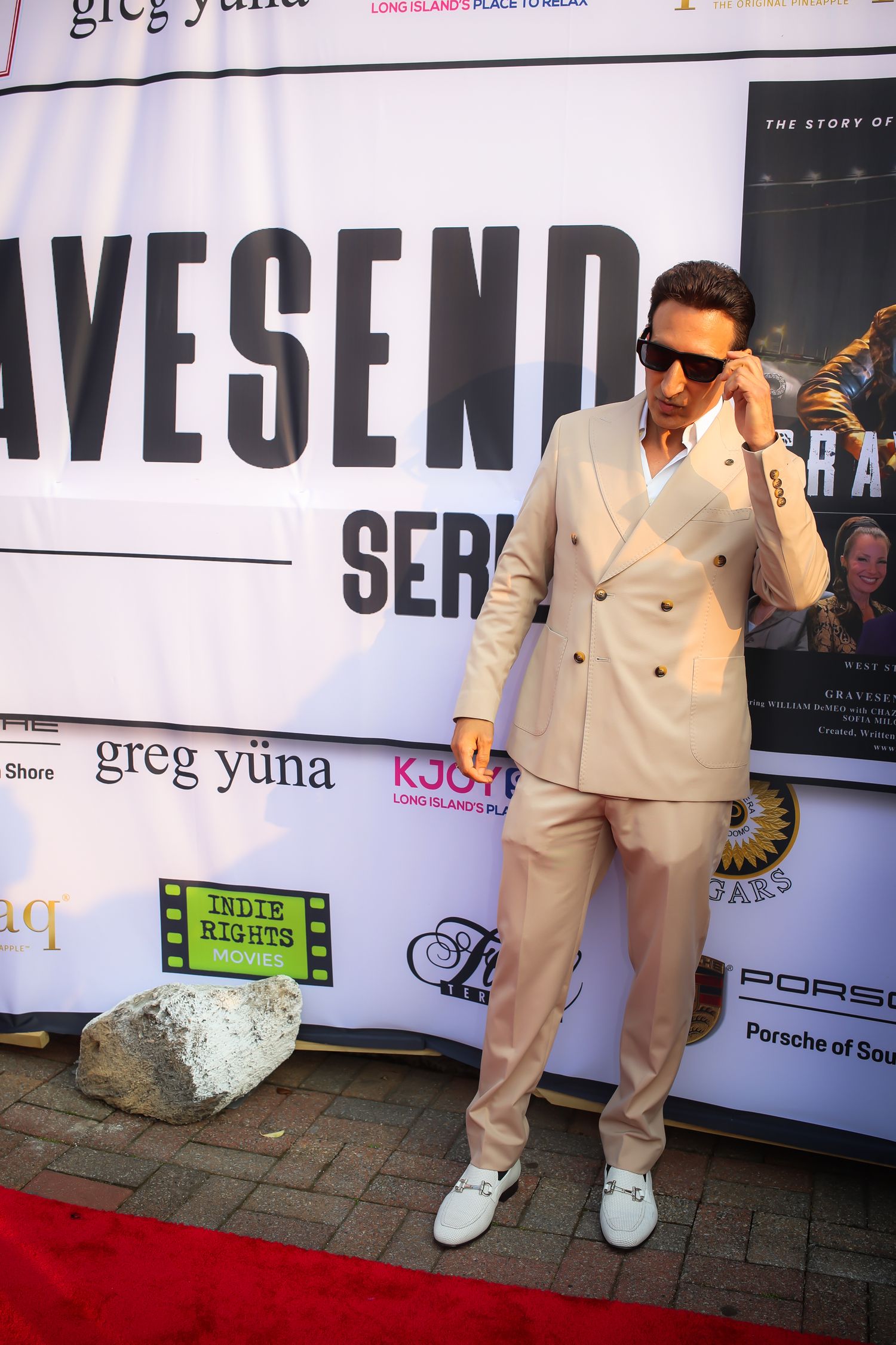 A person in a beige suit and white sneakers poses at different angles against a backdrop with text and logos.