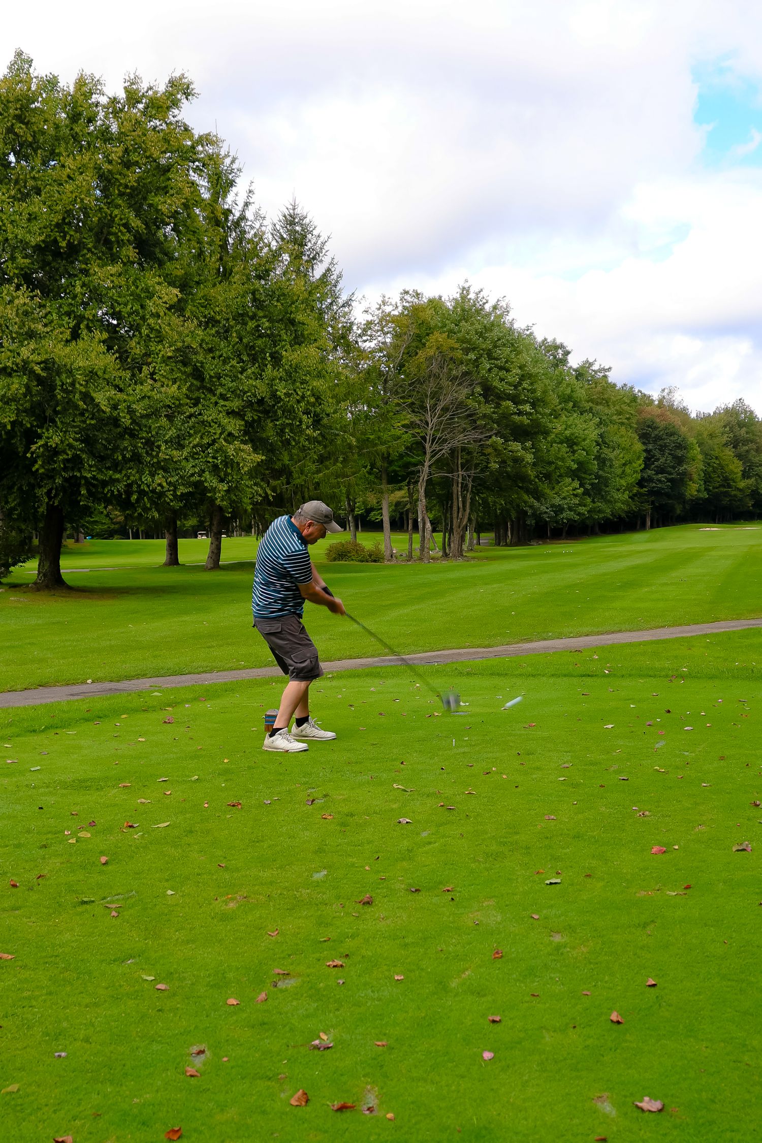 A golfer takes a swing on a lush green course surrounded by trees on a partly cloudy day.