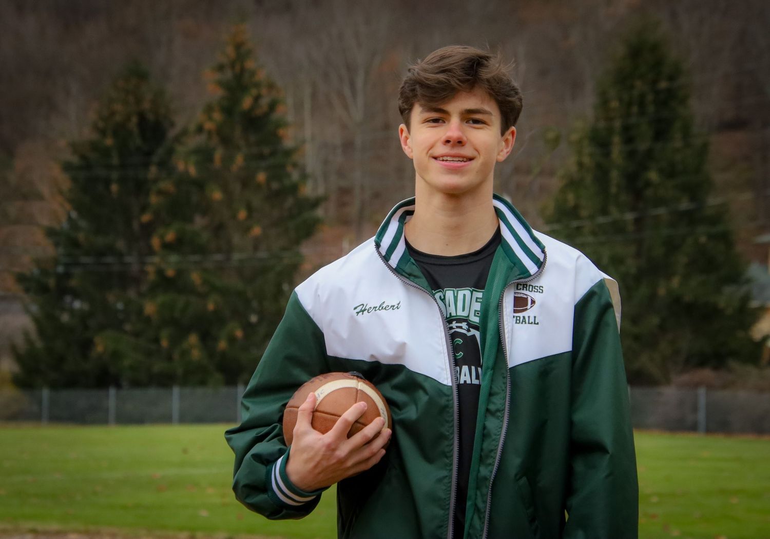 High school athlete in green and white uniform poses with football in outdoor sports field setting.