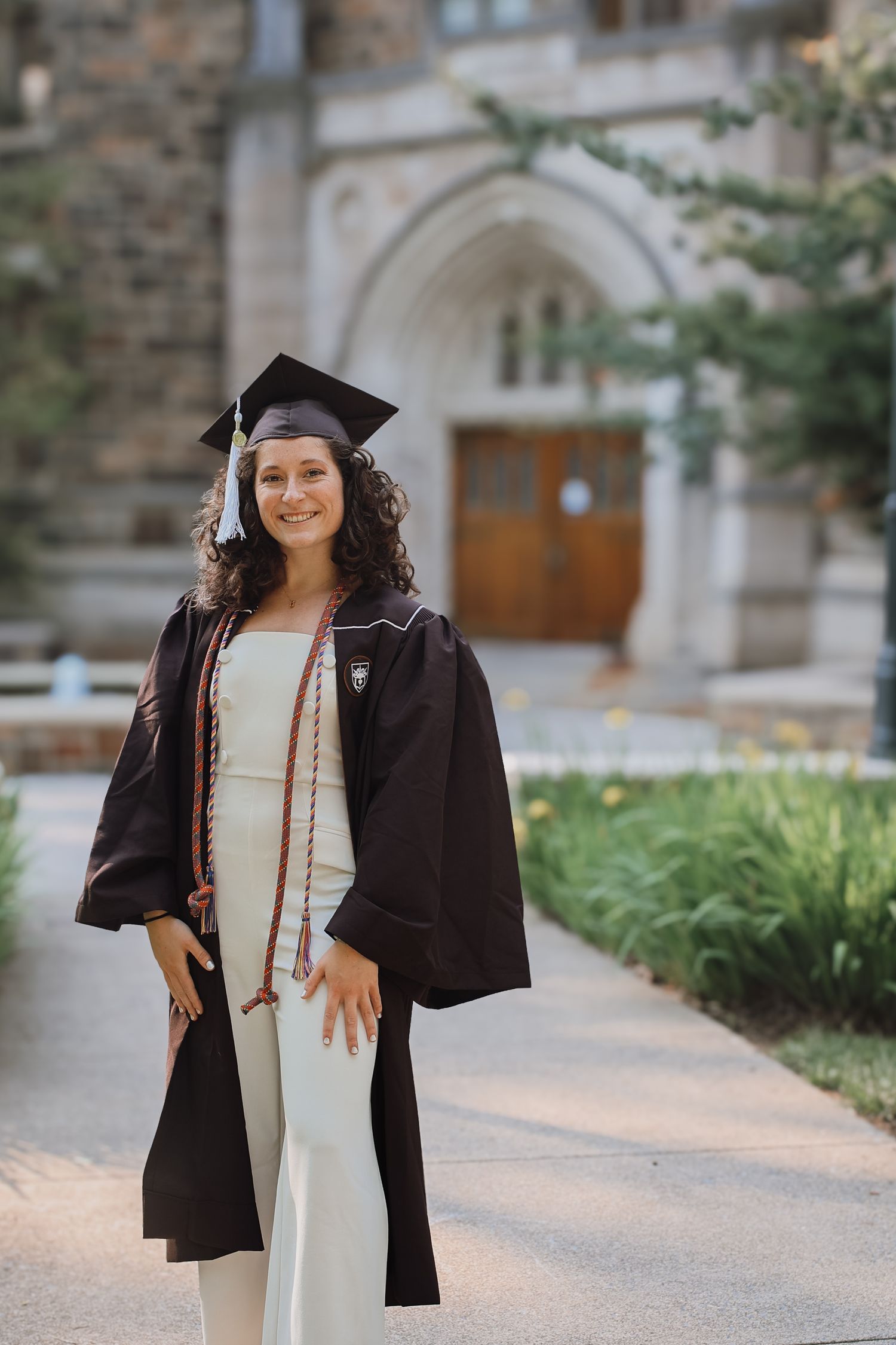 Graduate in black cap and gown poses for graduation photos on a stone path surrounded by greenery.