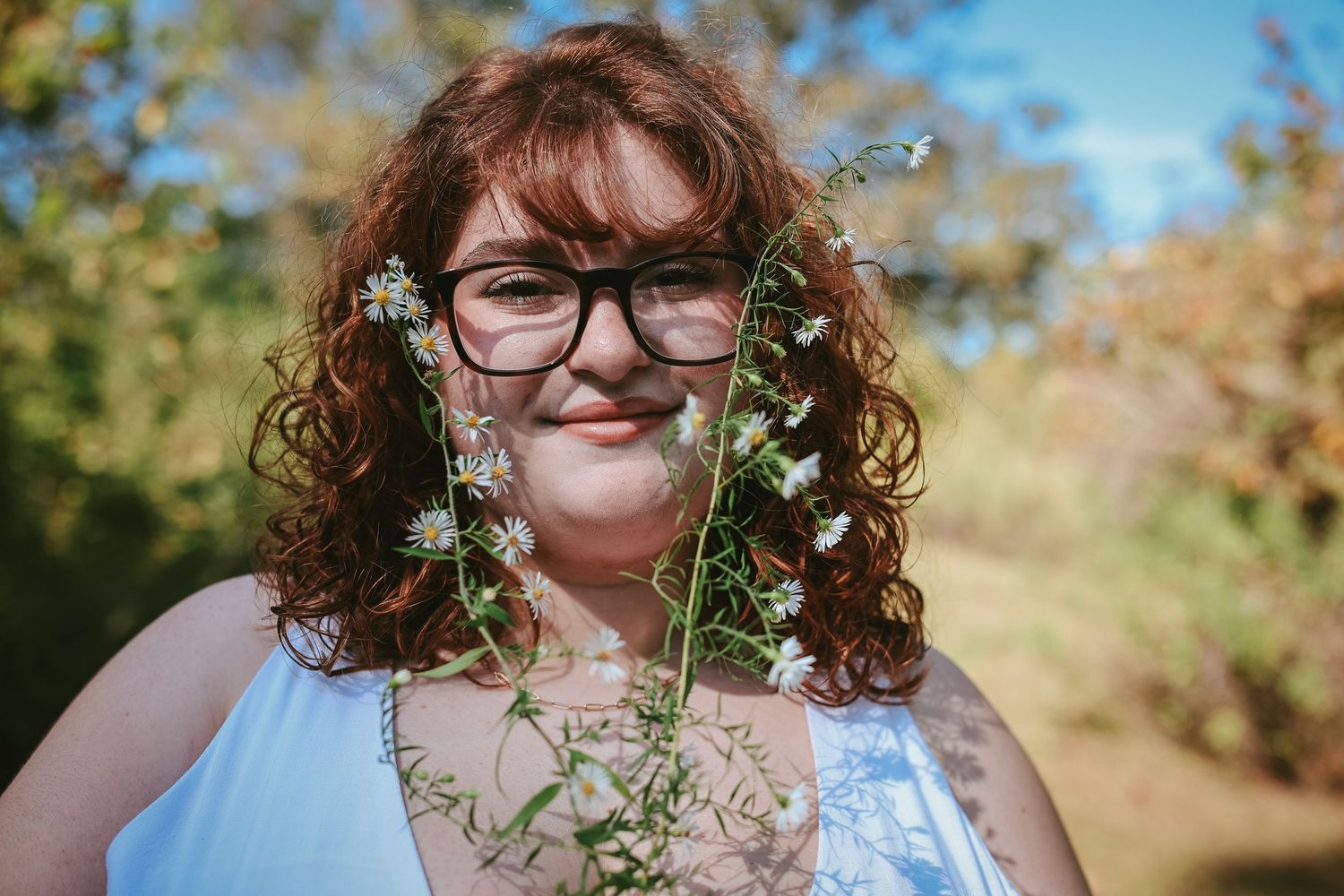 A person with curly red hair and glasses poses outdoors with delicate white flowers in a sunny garden setting.