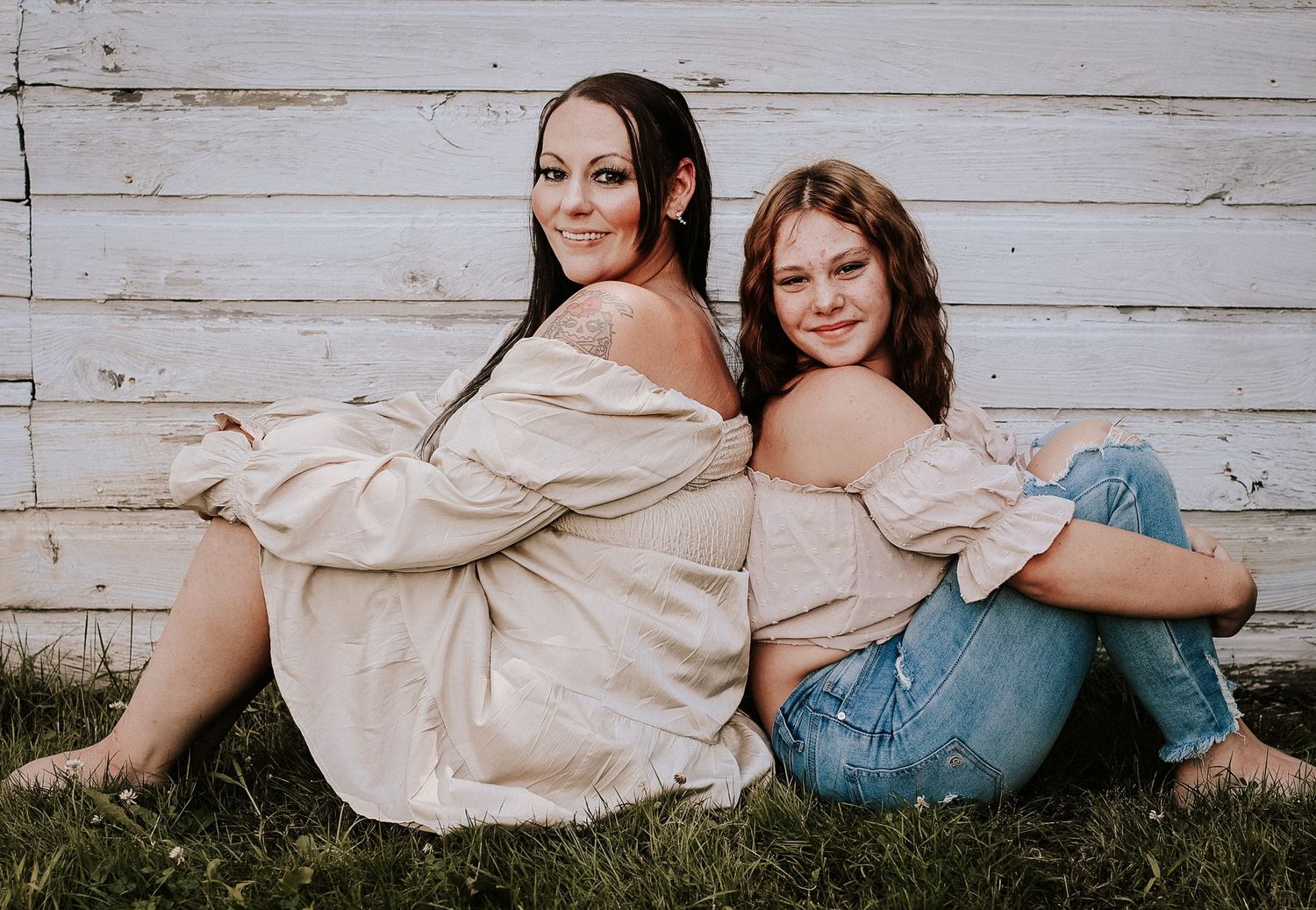Two people in matching beige off-shoulder tops and jeans sit back-to-back against rustic white wooden wall outdoors.