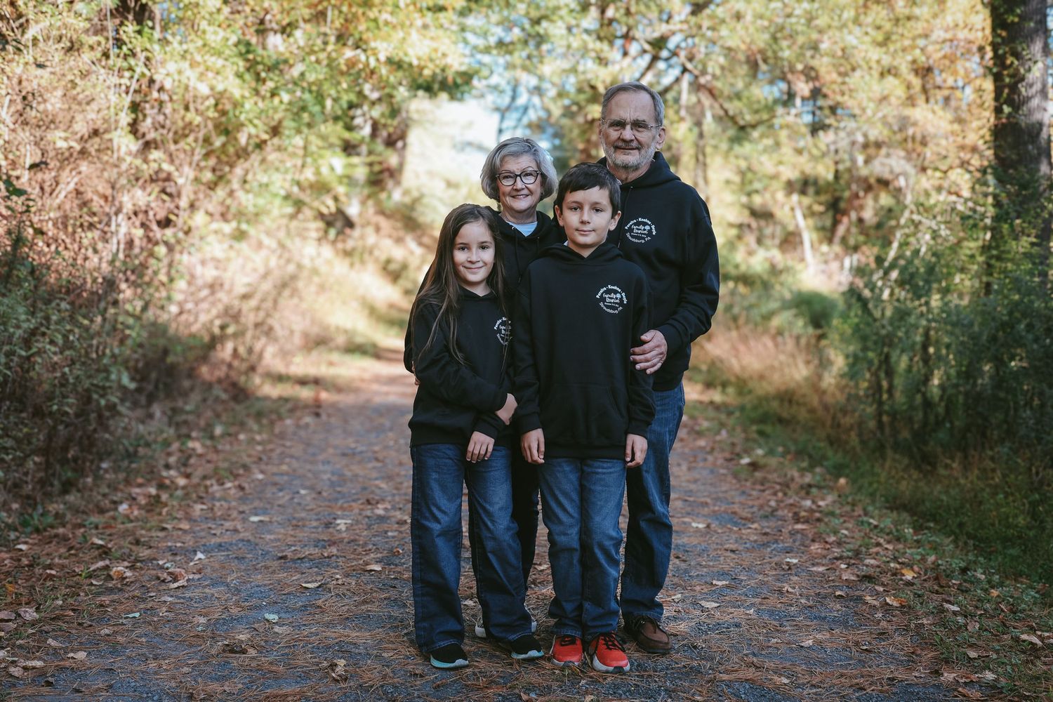 A group wearing matching black hoodies stands together on a leaf-covered forest path during autumn.