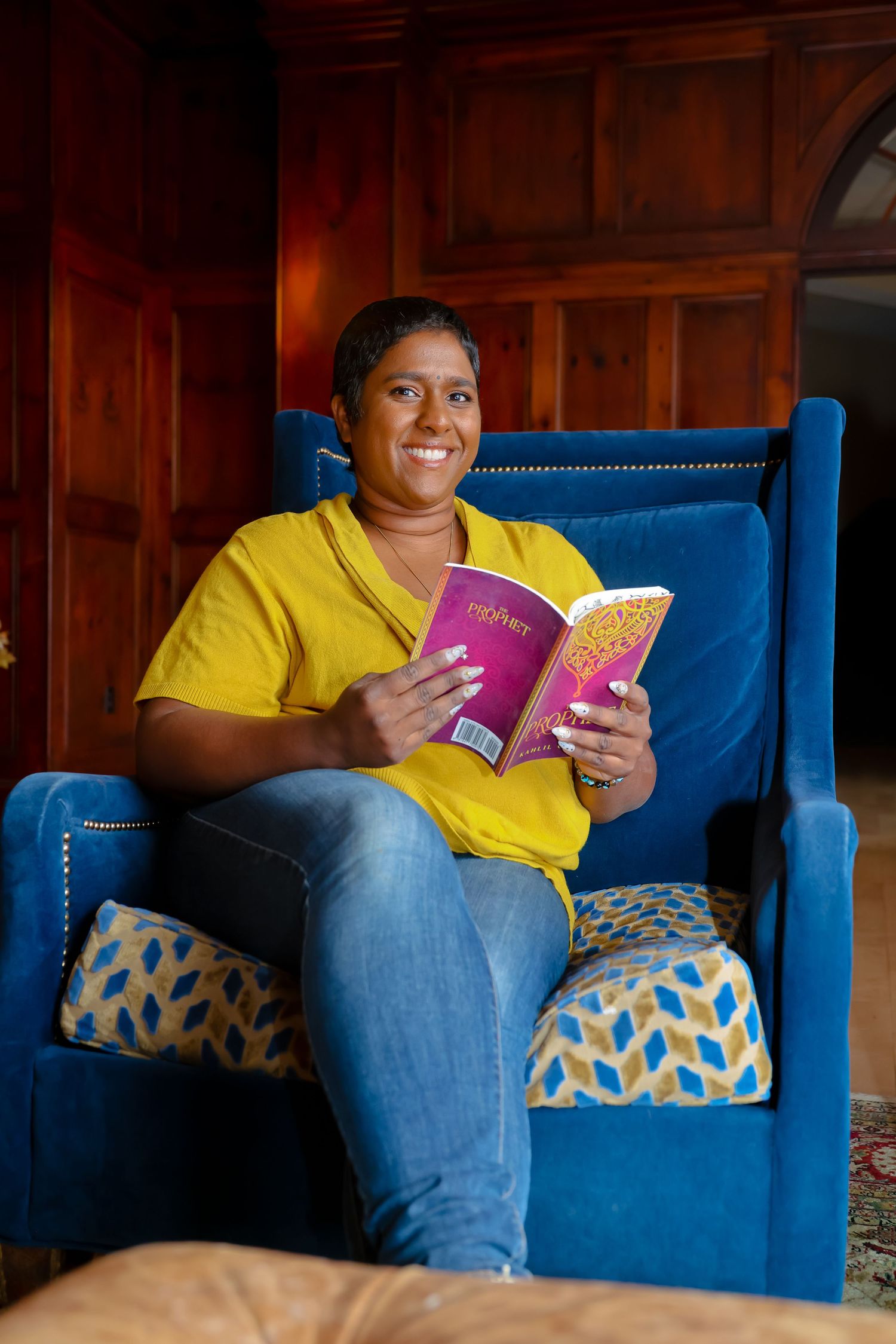 Someone relaxes on a bright blue armchair while reading a pink book in a wood-paneled room.