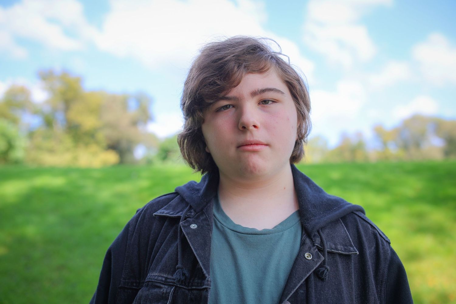 A person in a black jacket stands in a grassy field on a sunny day with blue skies and trees in the background.