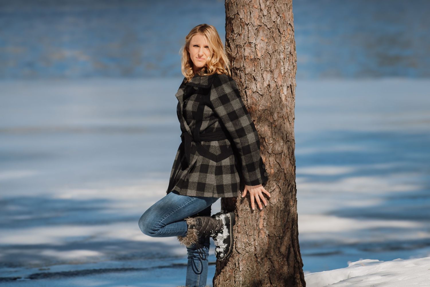 A person in a plaid coat and jeans poses against a tree trunk with snow and blue sky in the background.