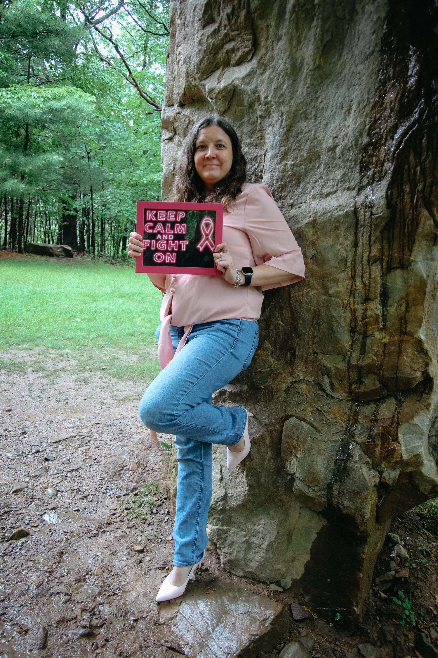 A person in jeans and a pink shirt sits on a rocky ledge in a wooded area holding a sign.