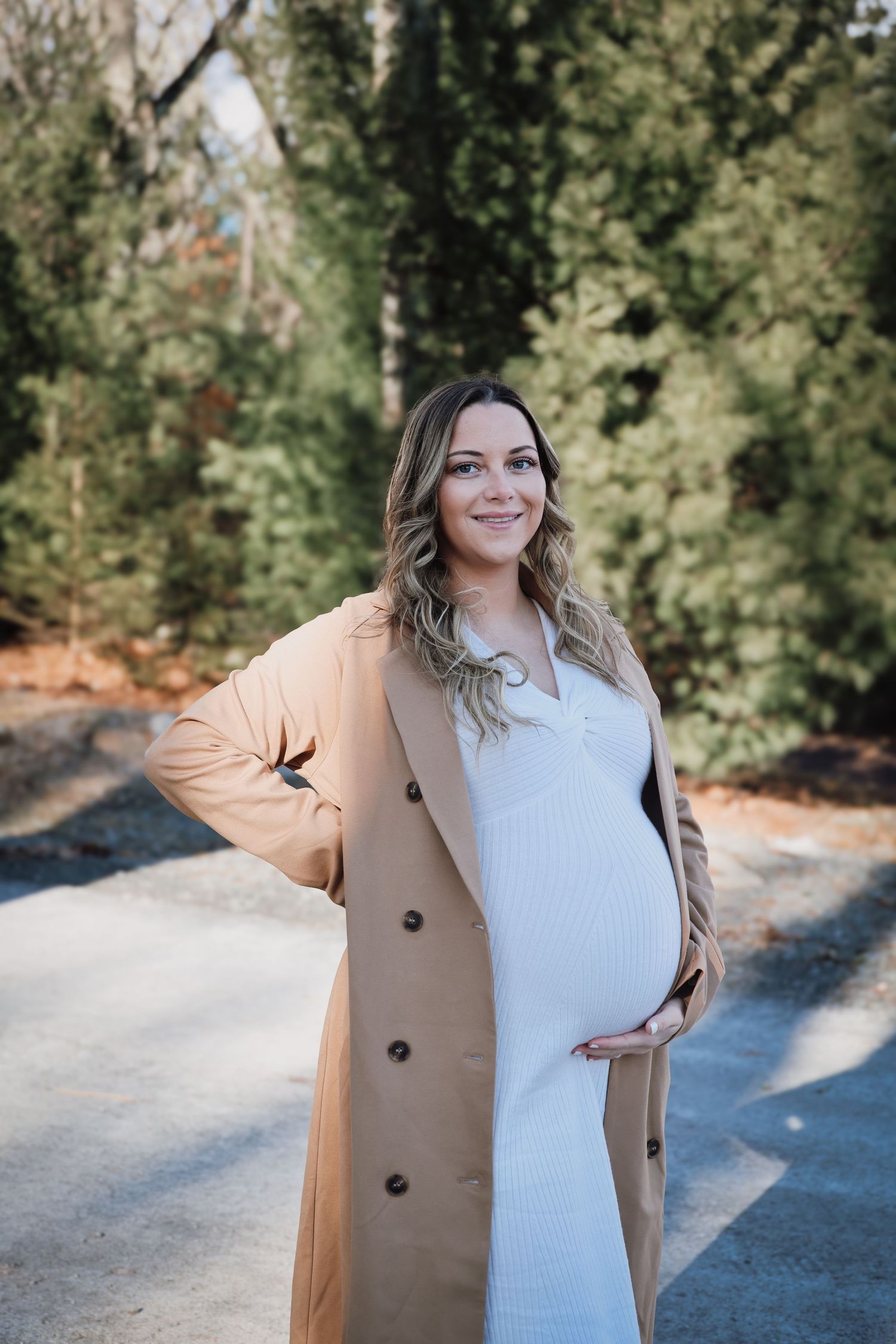 A pregnant woman in a white dress and tan trench coat stands on a path surrounded by evergreen trees.