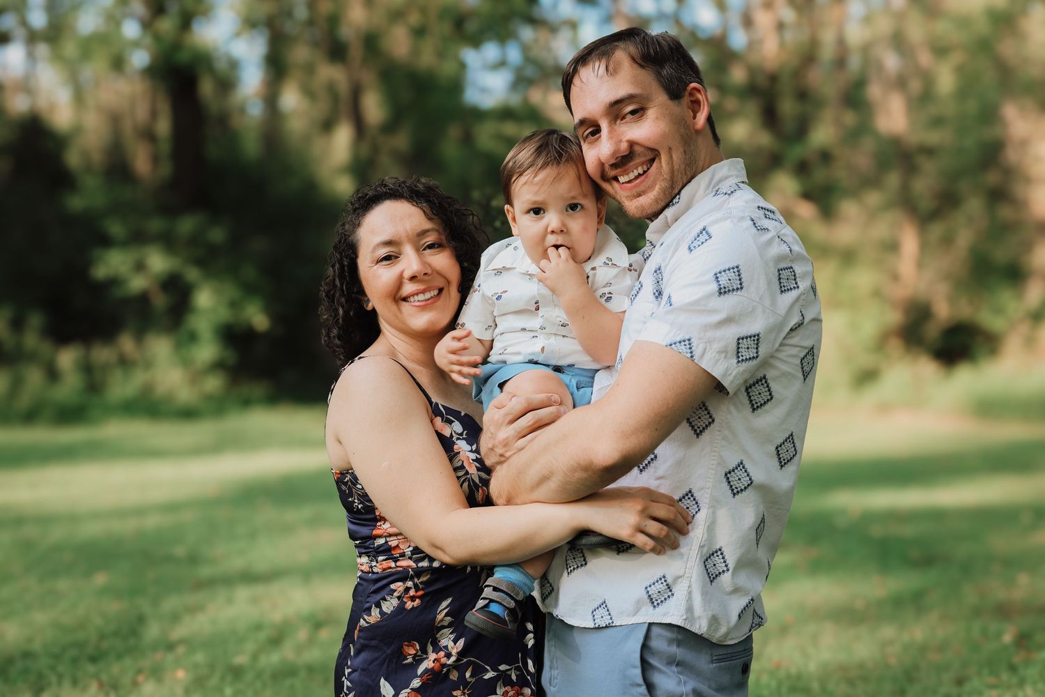 A family poses together outdoors on a sunny day in a grassy park setting.