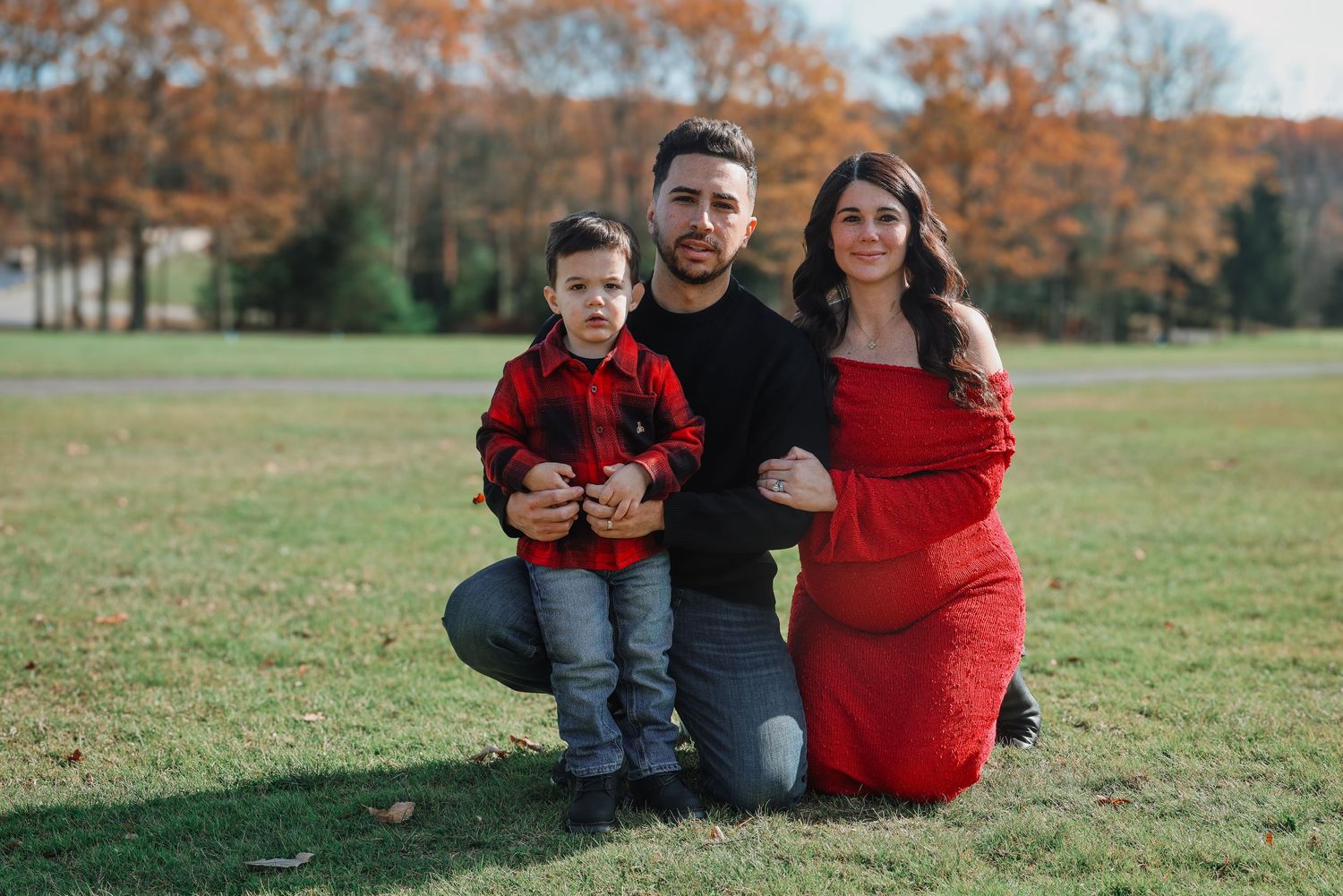 A family wearing red and black outfits poses together on grass during autumn with orange trees in background.