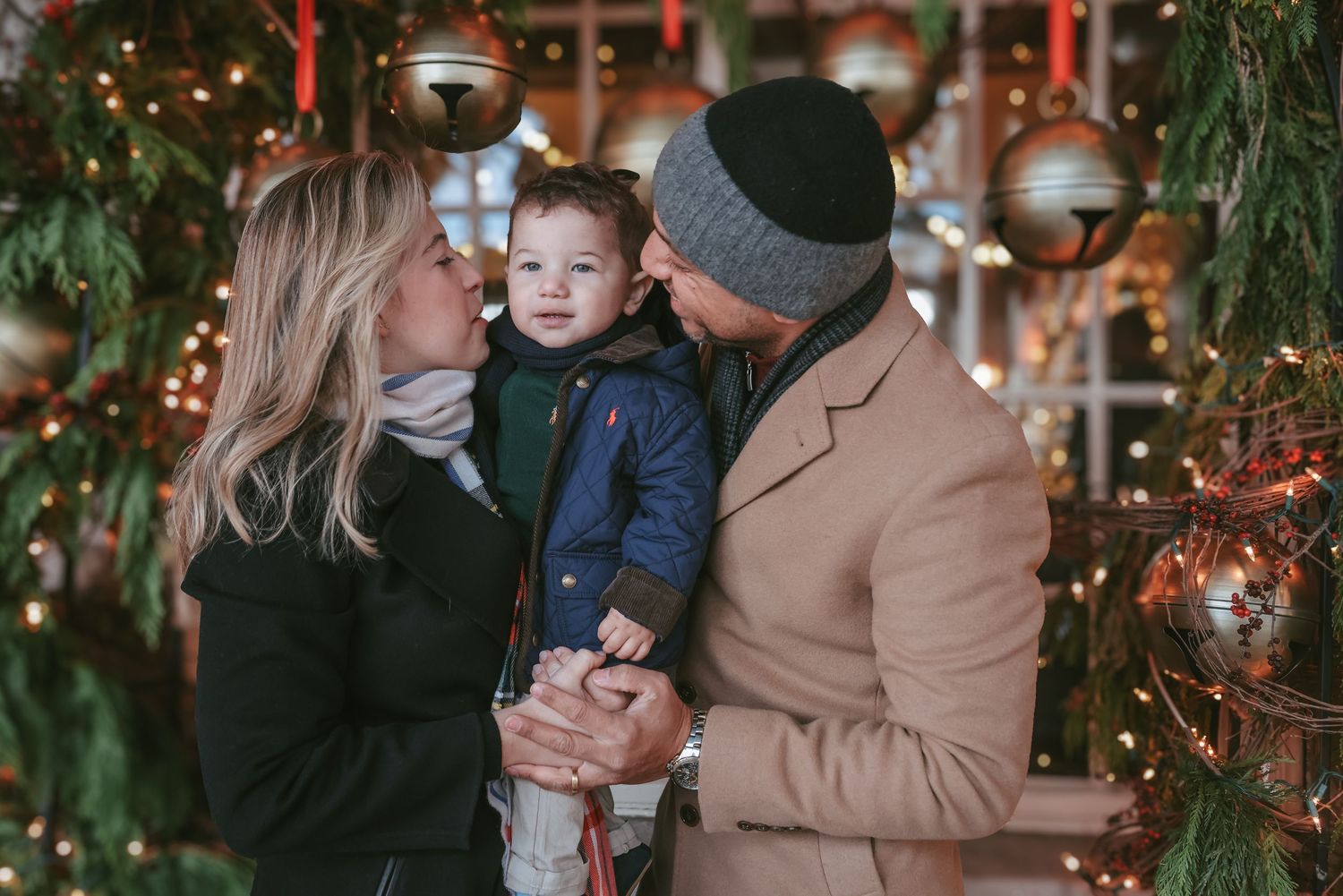 A family shares tender moments together during a Christmas photoshoot with festive lights and greenery in the background.