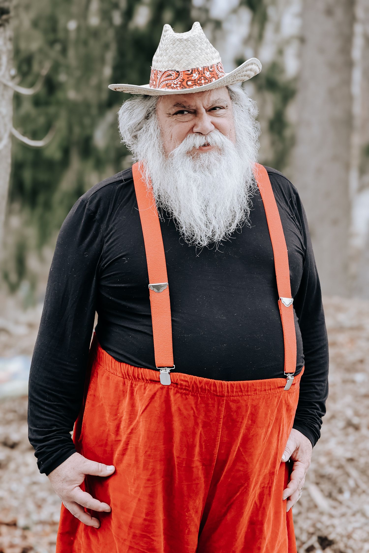 Person with long white beard wearing black shirt, orange suspenders and matching orange pants with a decorative hat.