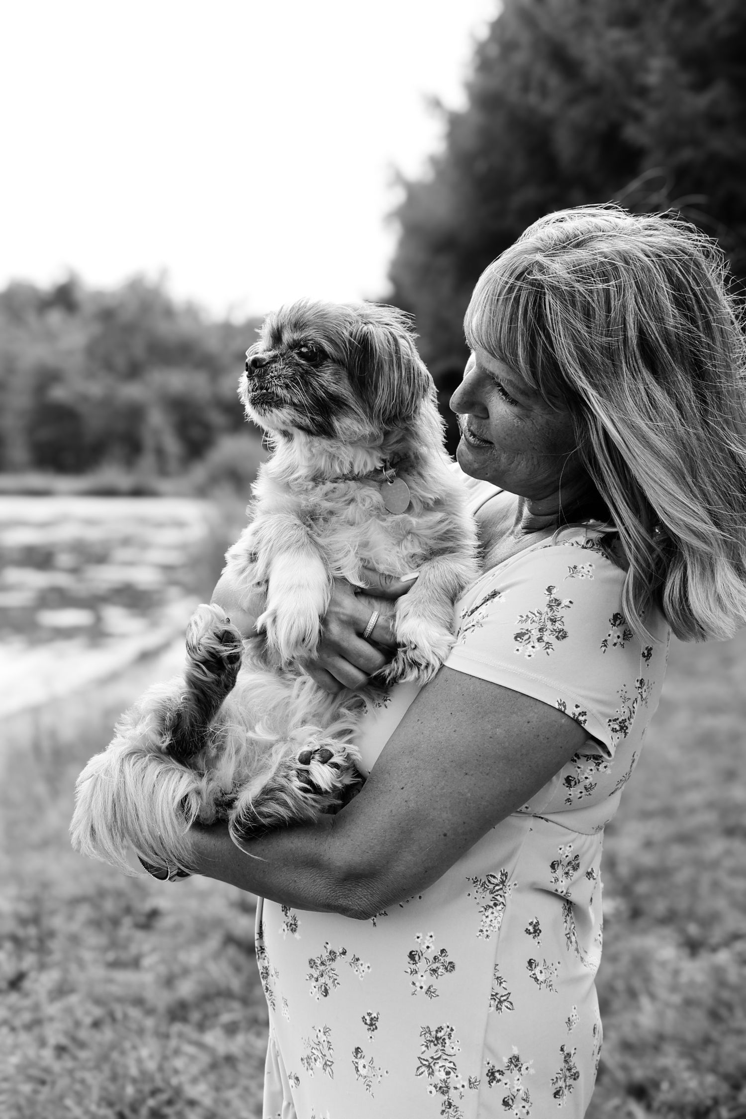 A person in a floral dress holds a small fluffy Shih Tzu dog outdoors in a black and white portrait.