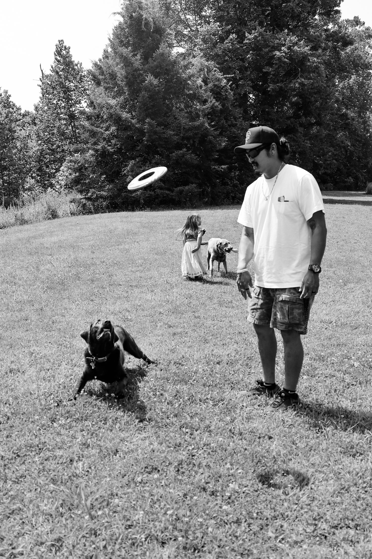 A person in casual attire plays frisbee with a black dog on a grassy lawn near trees in a black and white photo.