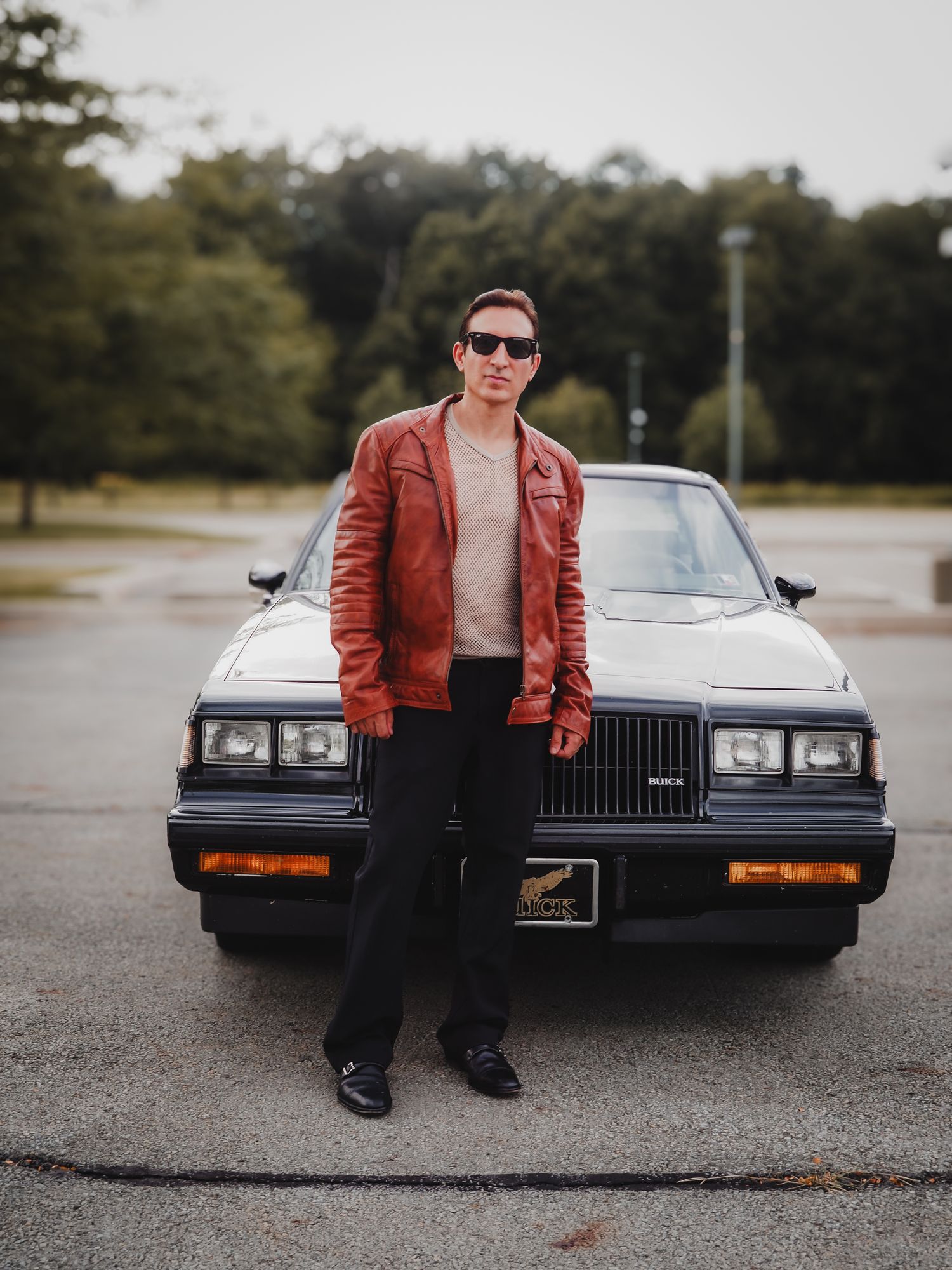 A vintage black Buick Grand National car parked in an empty lot with someone in a red leather jacket posing near it.