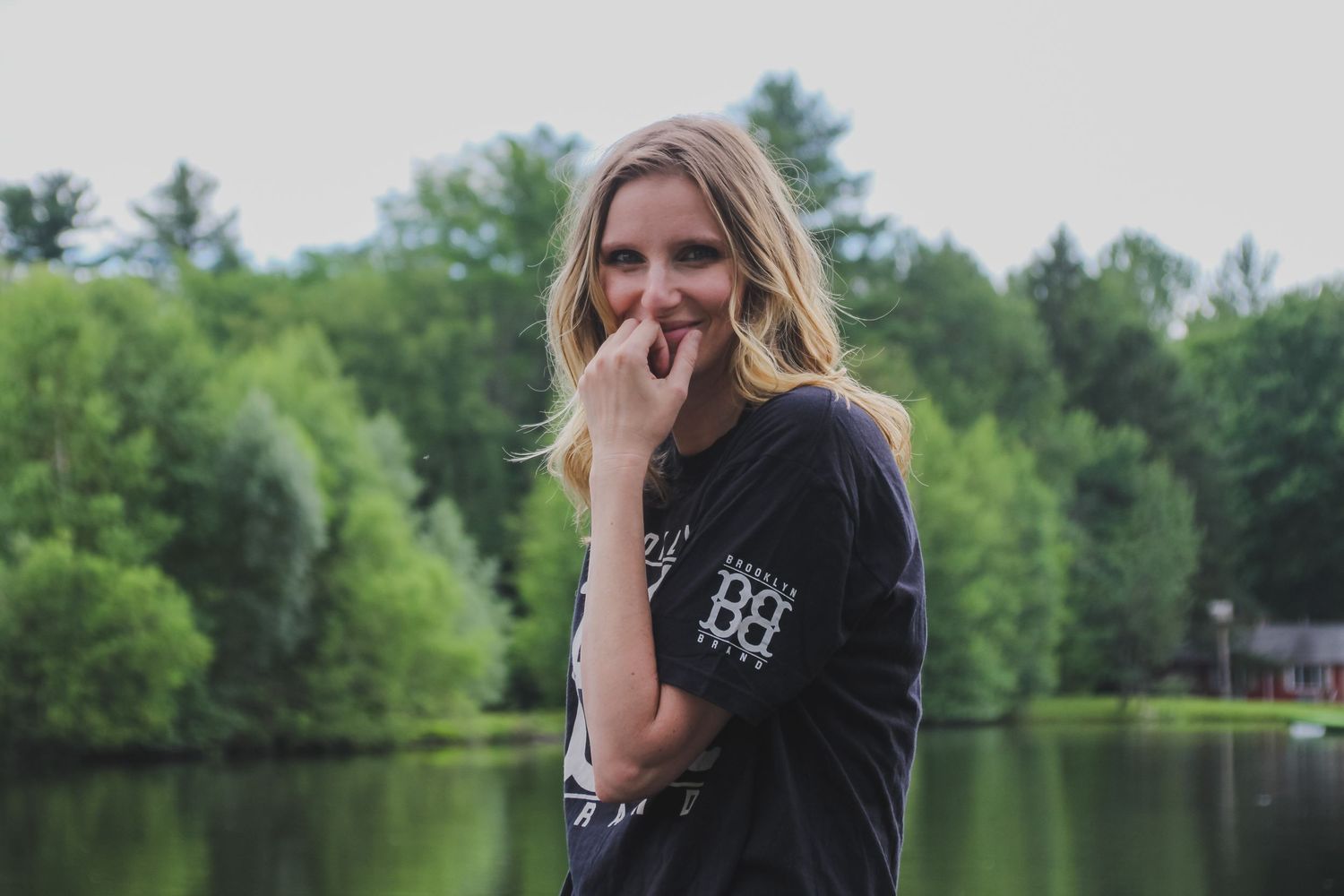 A person in a black shirt stands near a tranquil lake surrounded by lush green trees on a sunny day.