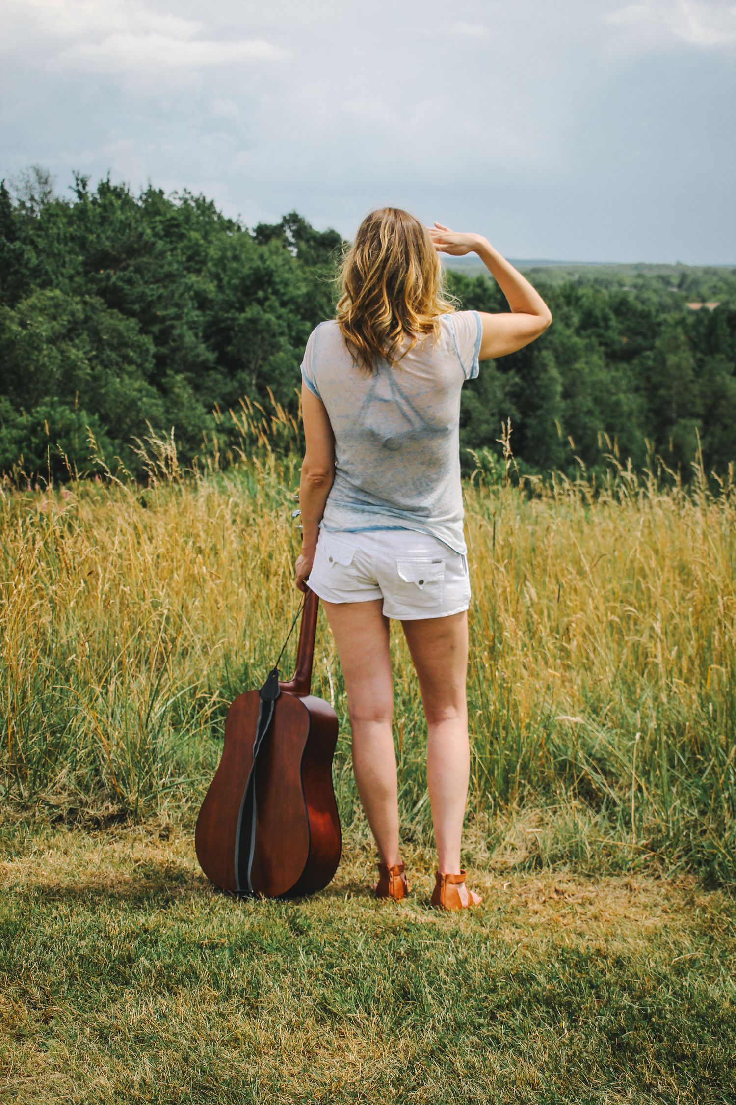 Acoustic guitar rests beside someone in white shorts sitting in a golden summer meadow overlooking a scenic landscape.