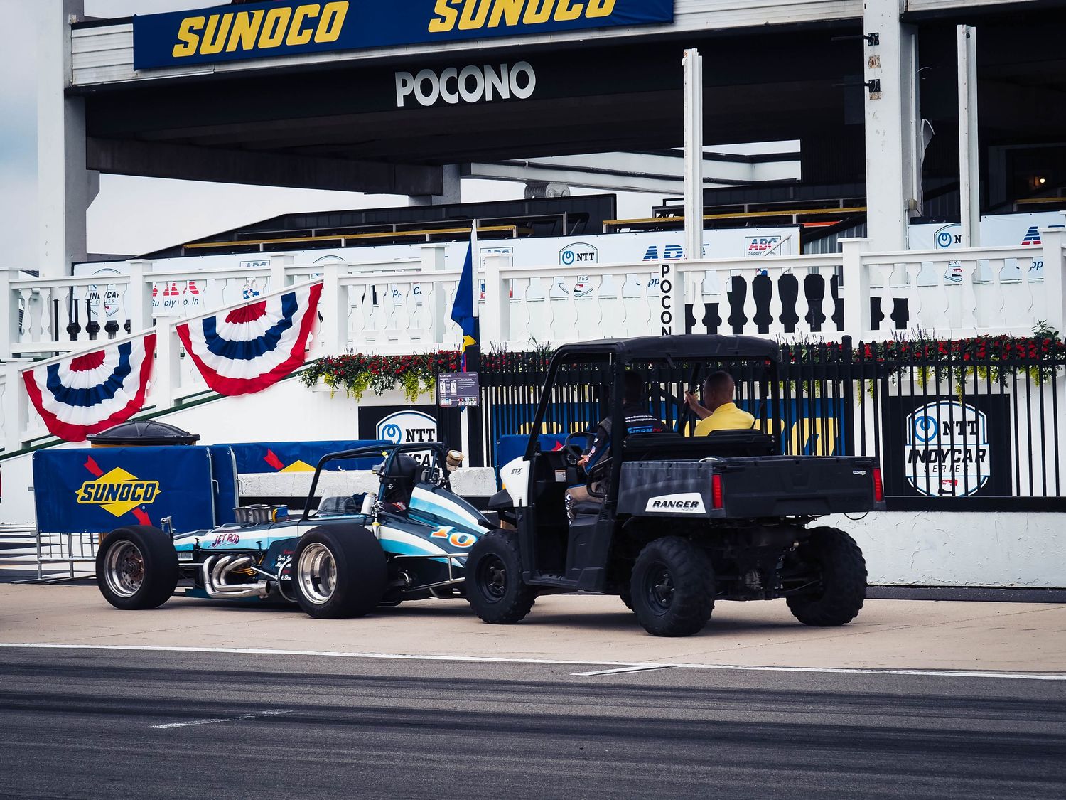 Race cars line up at a Sunoco-branded pit lane with American flag bunting decorating the grandstands at a motorsport track.