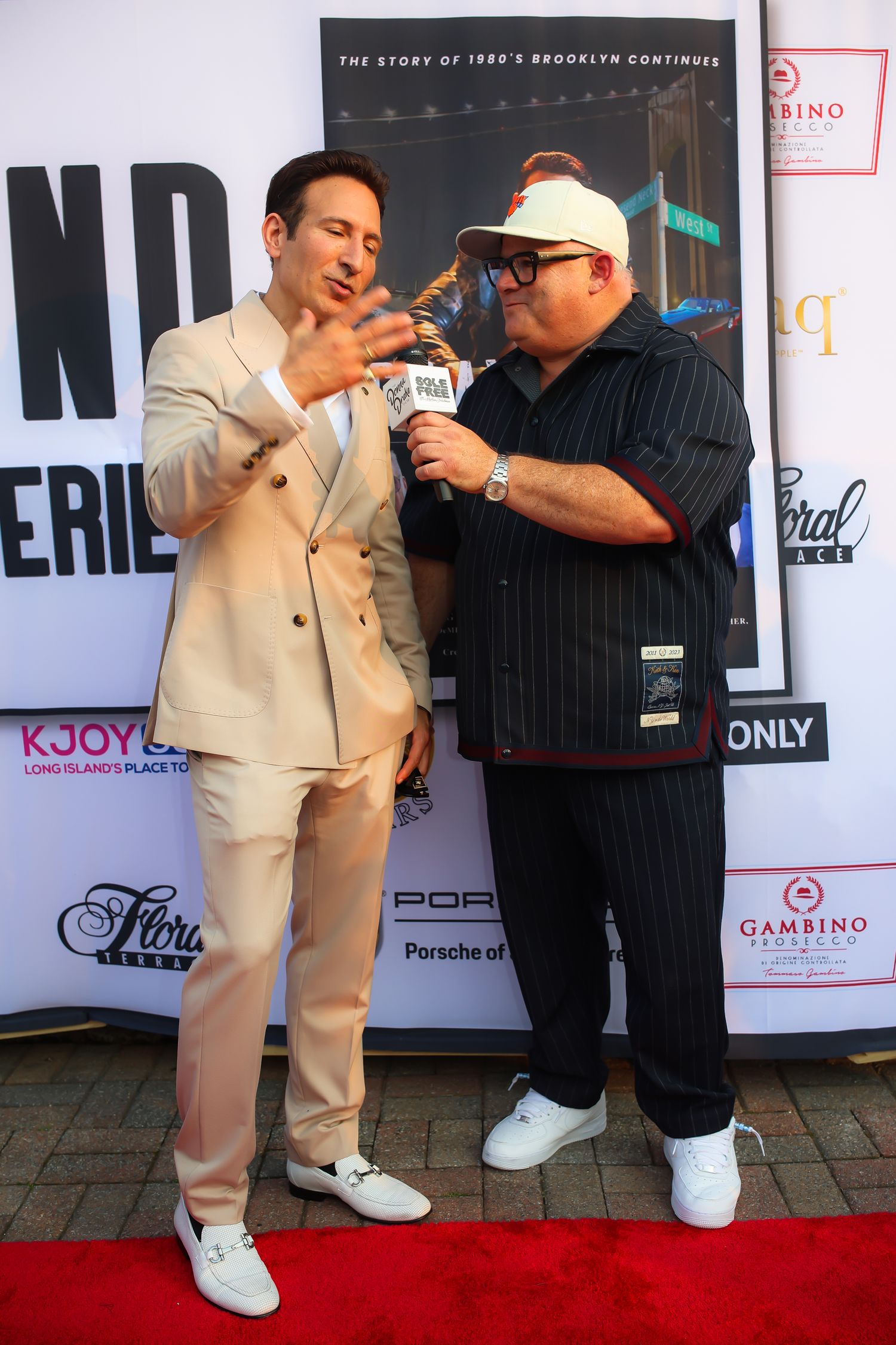 Two people pose together on a red carpet event, one wearing a beige suit and the other in black casual attire.