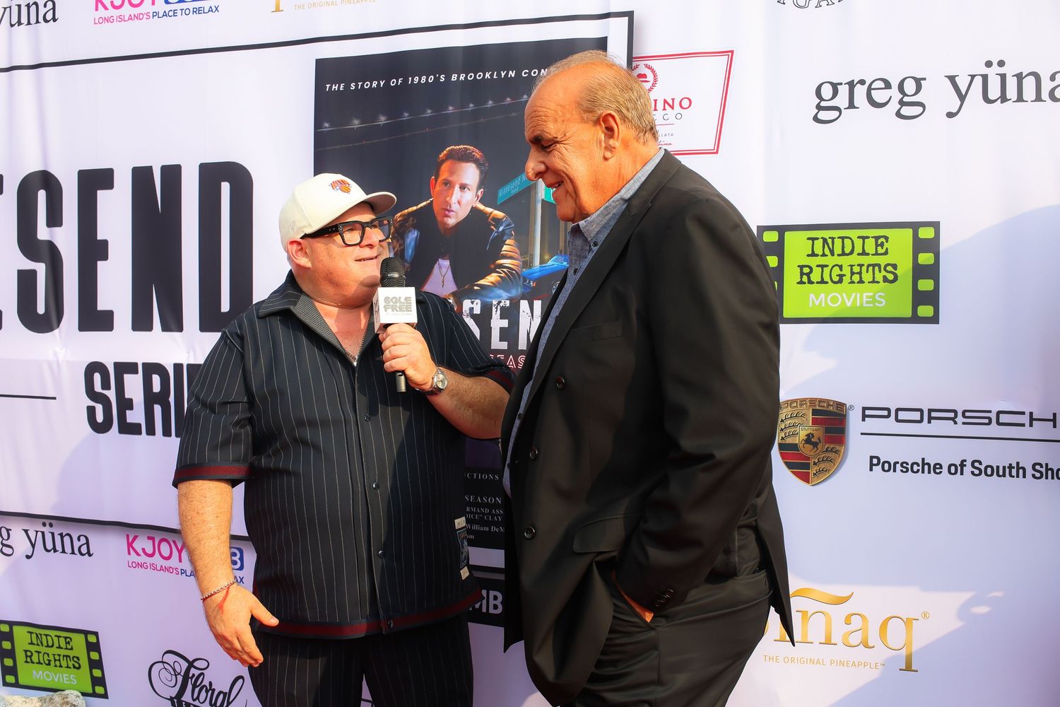People in formal attire stand against a backdrop with sponsor logos at a red carpet event.