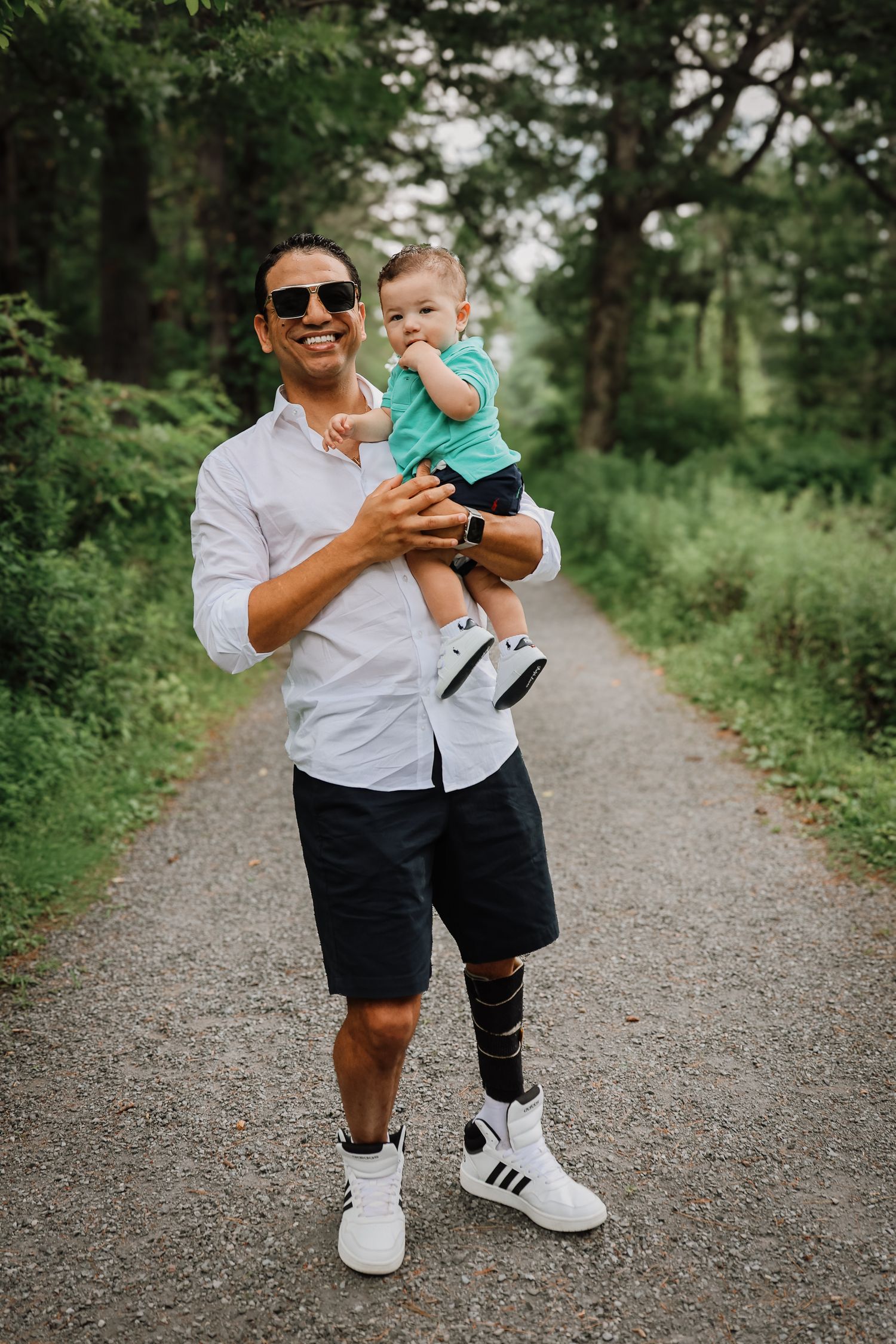 A pair walks together on a gravel path surrounded by lush green trees and vegetation on a sunny day.