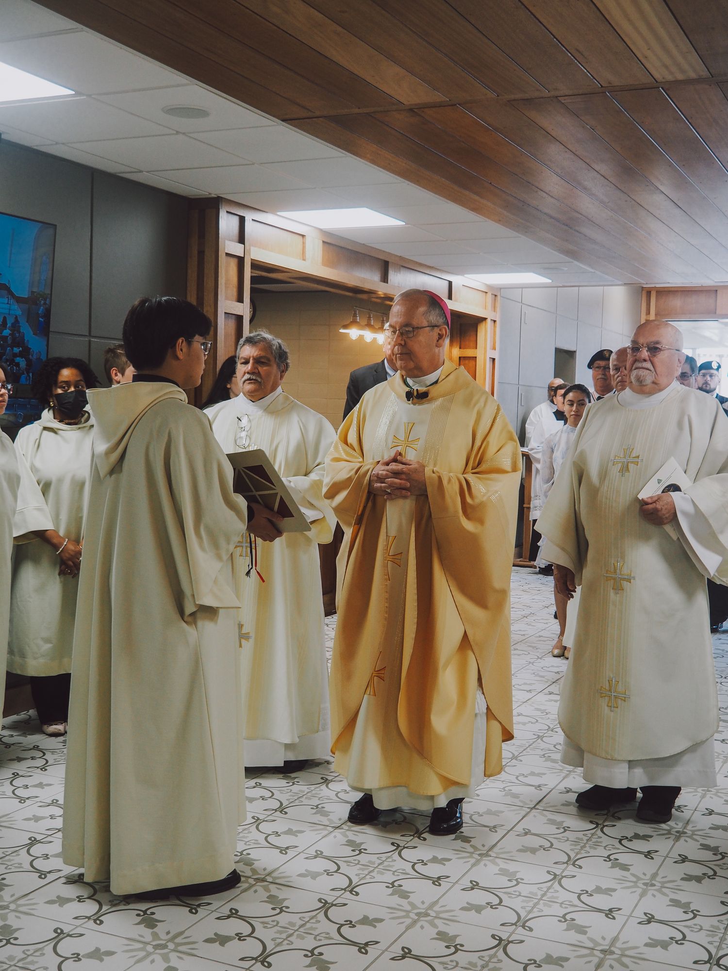 A group of clergy members in white and gold robes gather in a hallway with wooden ceiling panels.