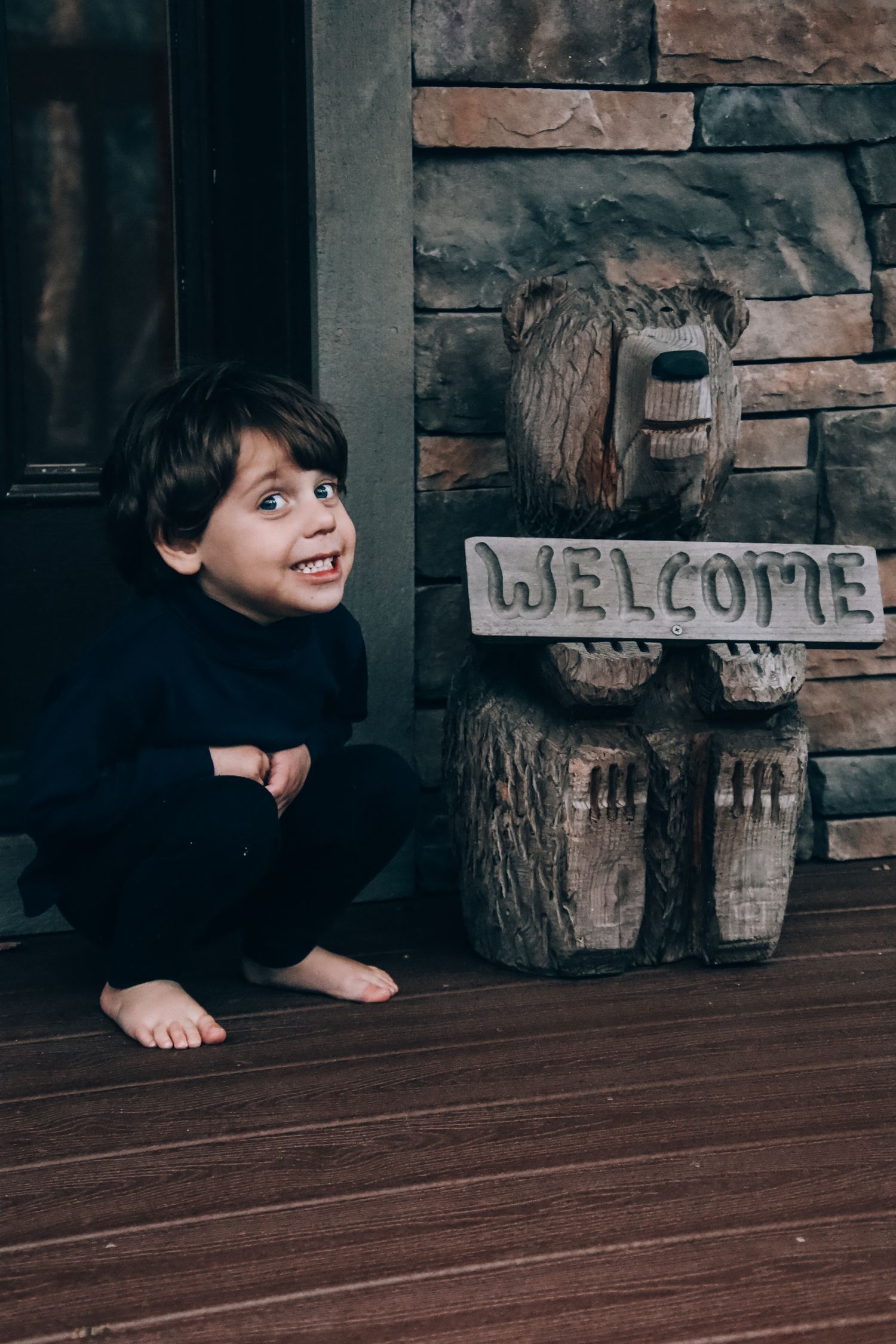 A child crouches next to a rustic wooden welcome bear carving on a dark wooden porch with stone wall backdrop.