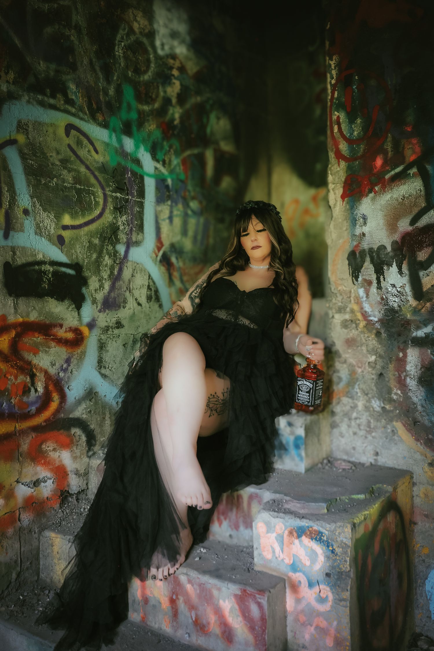 Figure in flowing black dress poses dramatically against graffiti-covered walls in abandoned urban setting.