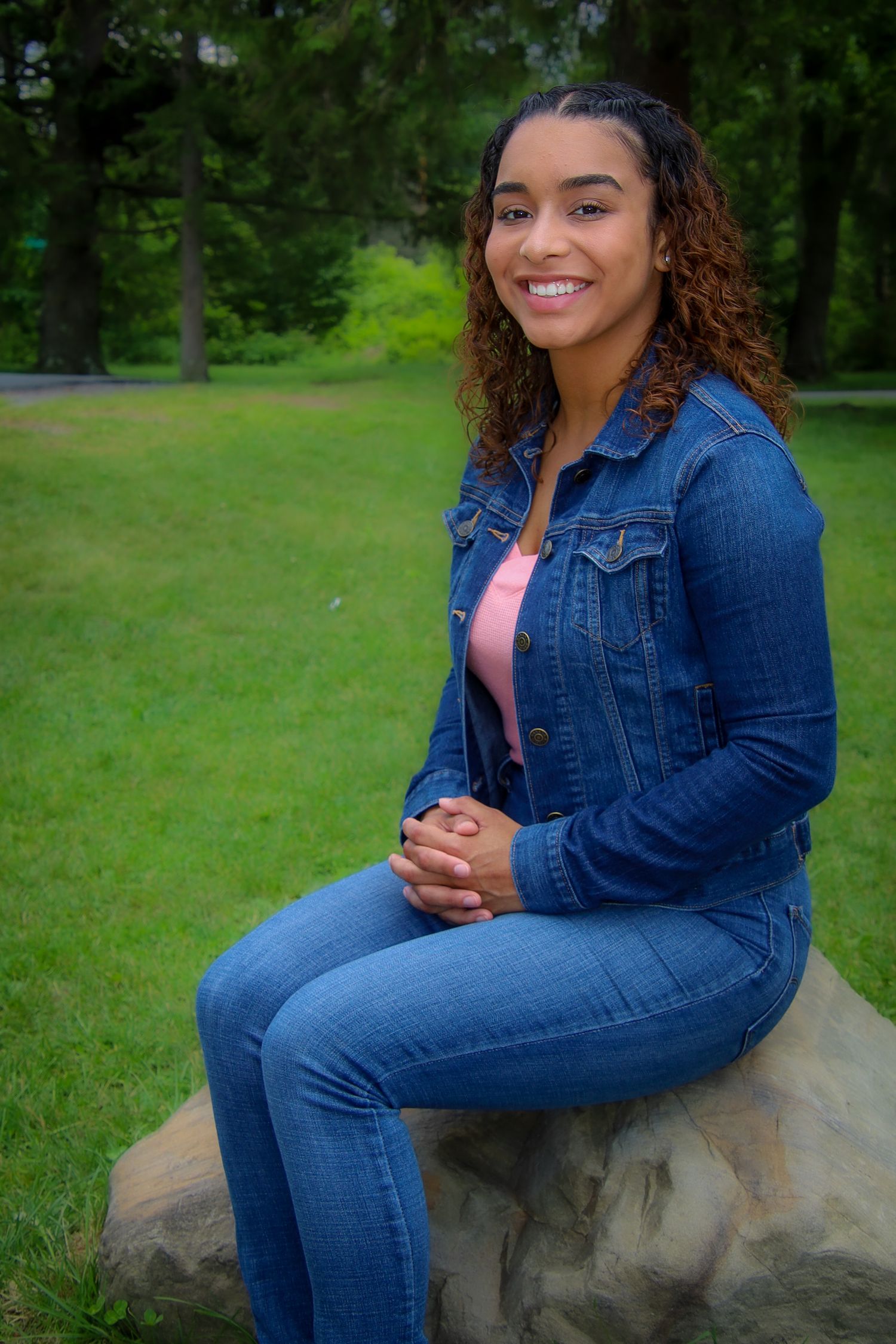 A young woman in denim jacket and jeans sits casually on a rock outdoors with green foliage in the background.