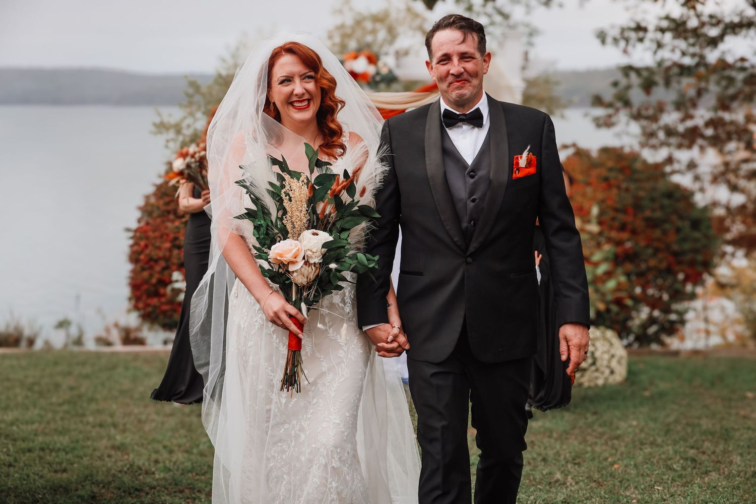 A couple in wedding attire walk together outdoors during autumn with colorful fall foliage in the background.