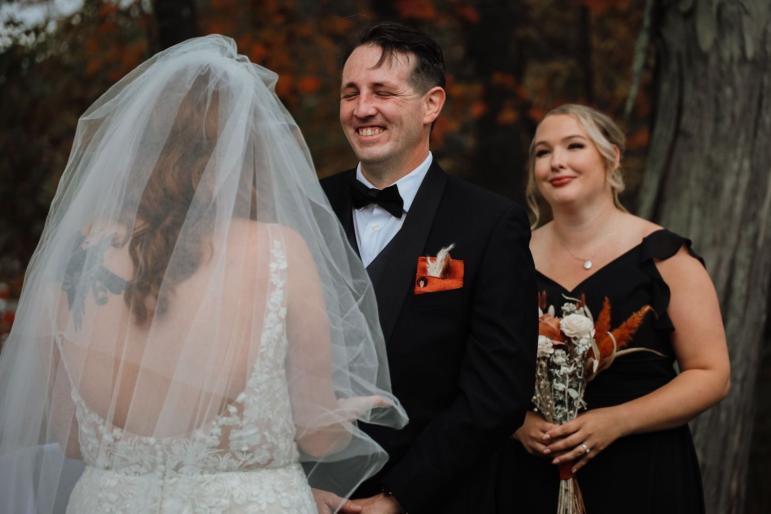 Wedding party members share a joyful moment during an autumn outdoor ceremony with fall foliage in background.