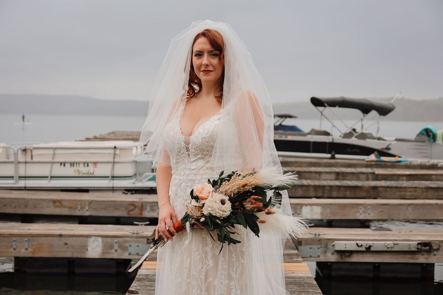 A bride in a white lace wedding dress holds a bouquet of white flowers on a wooden dock by the water.