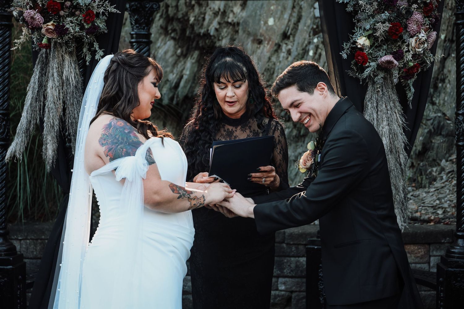 A couple exchanges rings during an outdoor wedding ceremony with an officiant under floral decorations and a rustic backdrop.