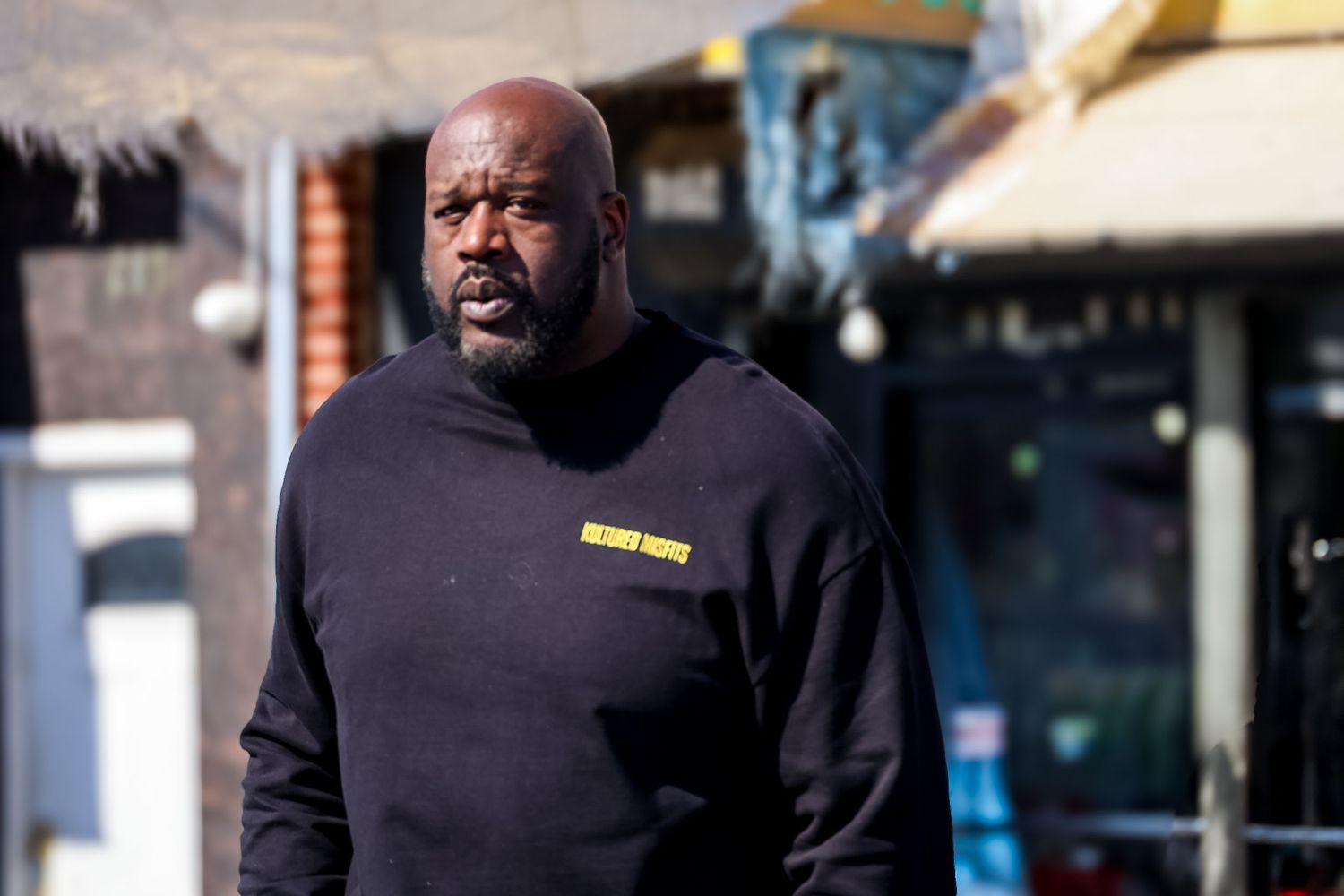 A person wearing a black sweater stands outside on a sunny day near storefronts.