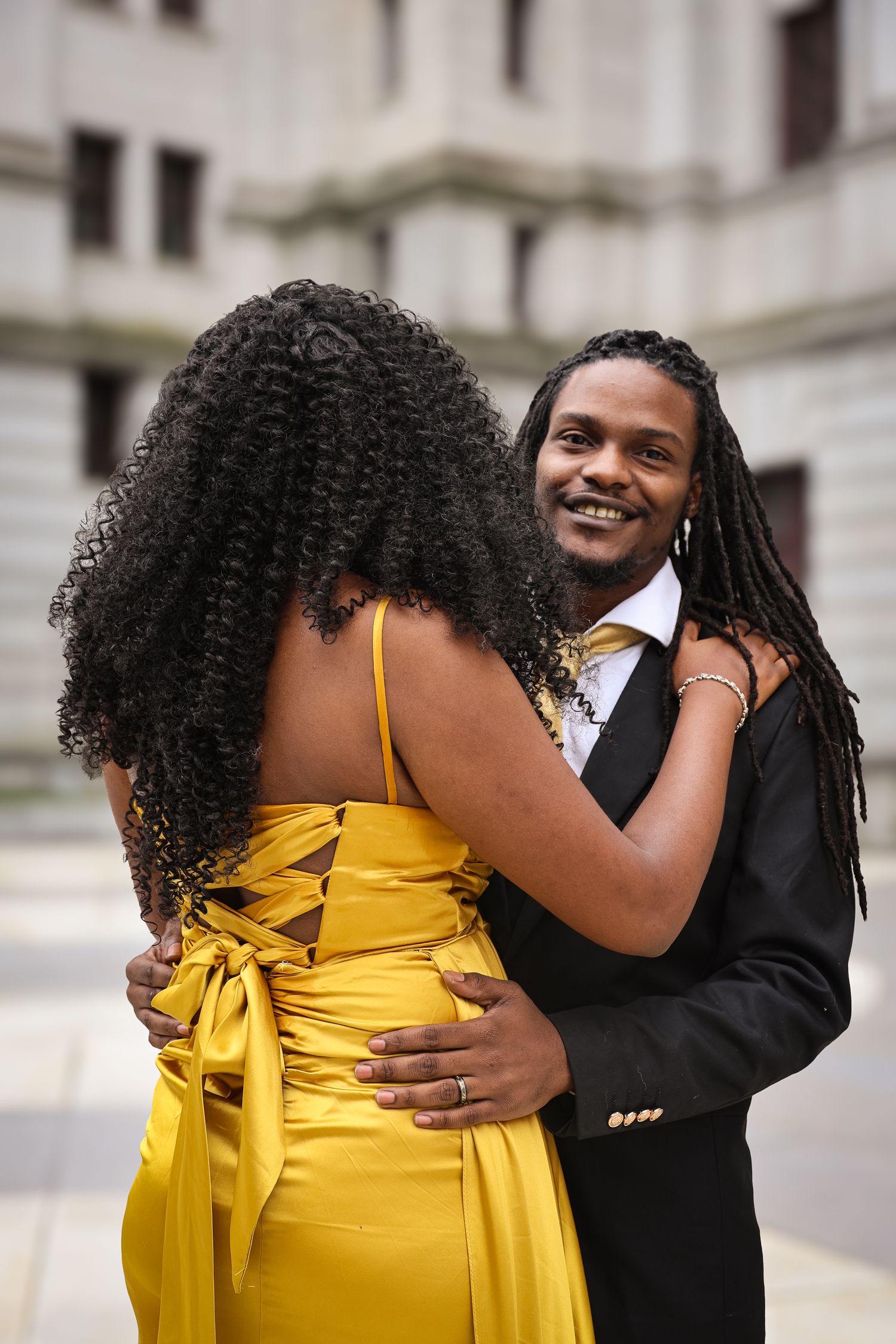 A couple in formal attire share an intimate moment, with one wearing a bright yellow dress and the other in a black suit.