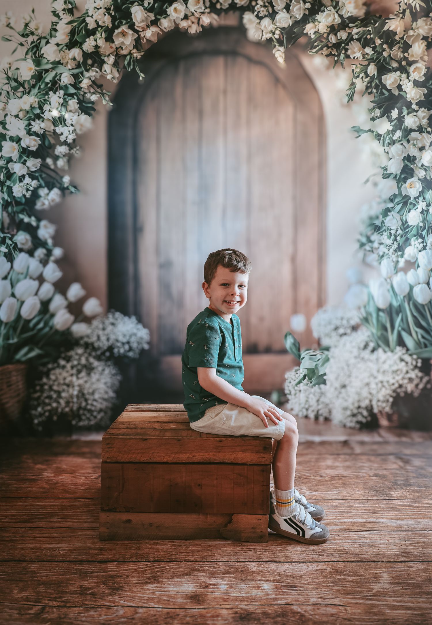 A small child sits on wooden steps surrounded by delicate white flowers and greenery in a rustic indoor setting.