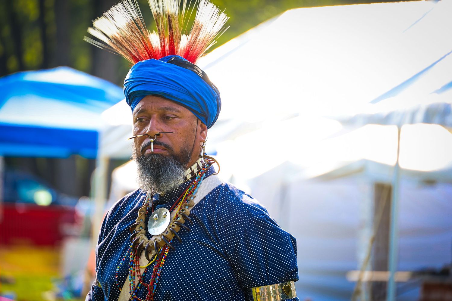 A person wearing traditional Native American regalia with a blue headdress and silver jewelry at an outdoor festival.