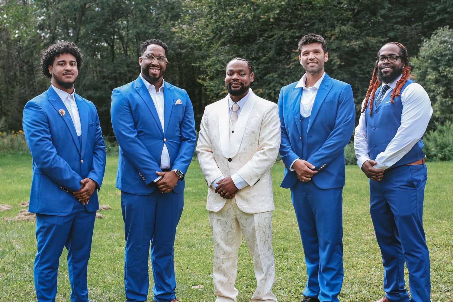 Wedding party wearing bright royal blue suits and one person in white pose together outdoors in a garden setting.