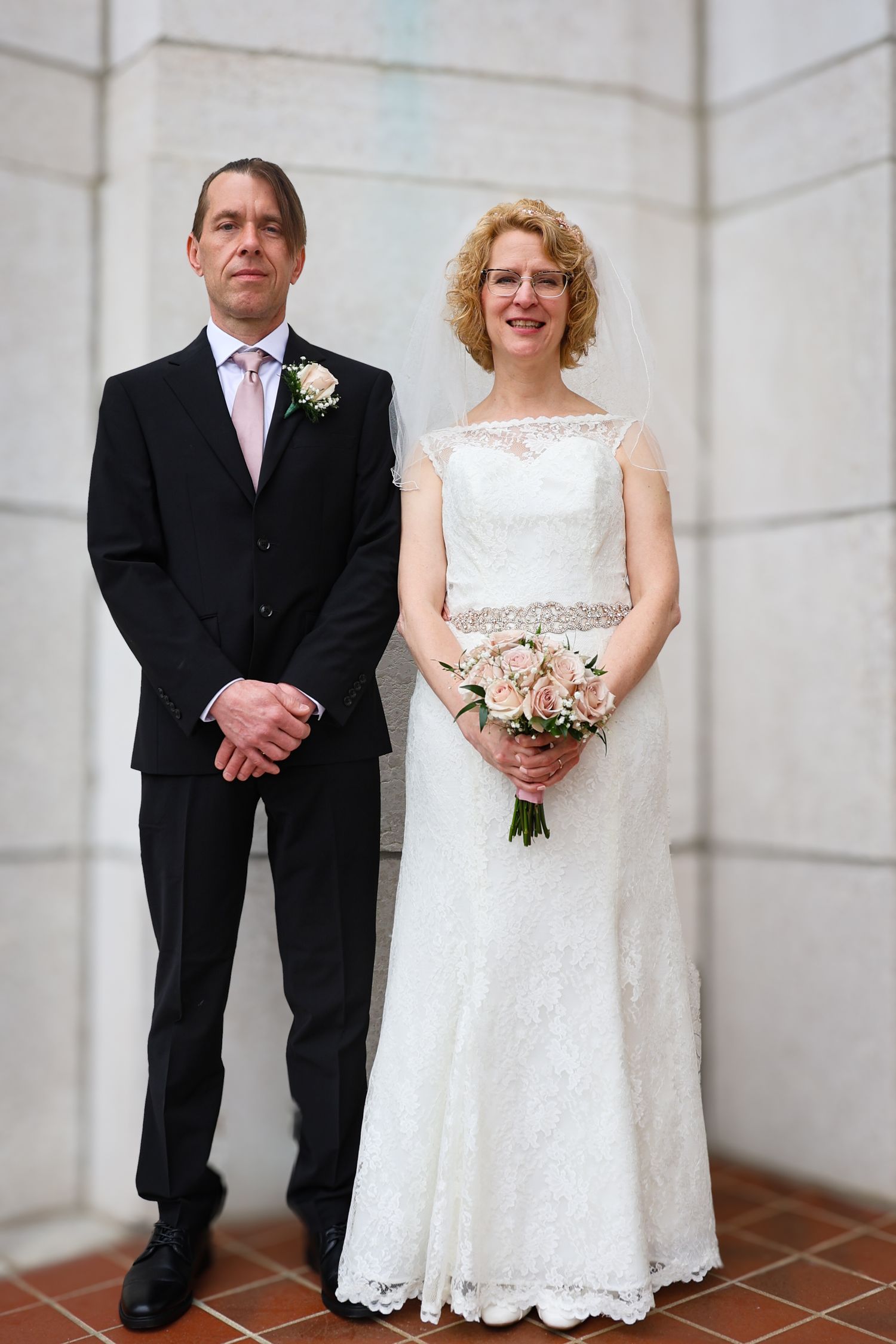 A wedding couple stands together in formal attire against a white corner wall, with the bride holding a pink bouquet.