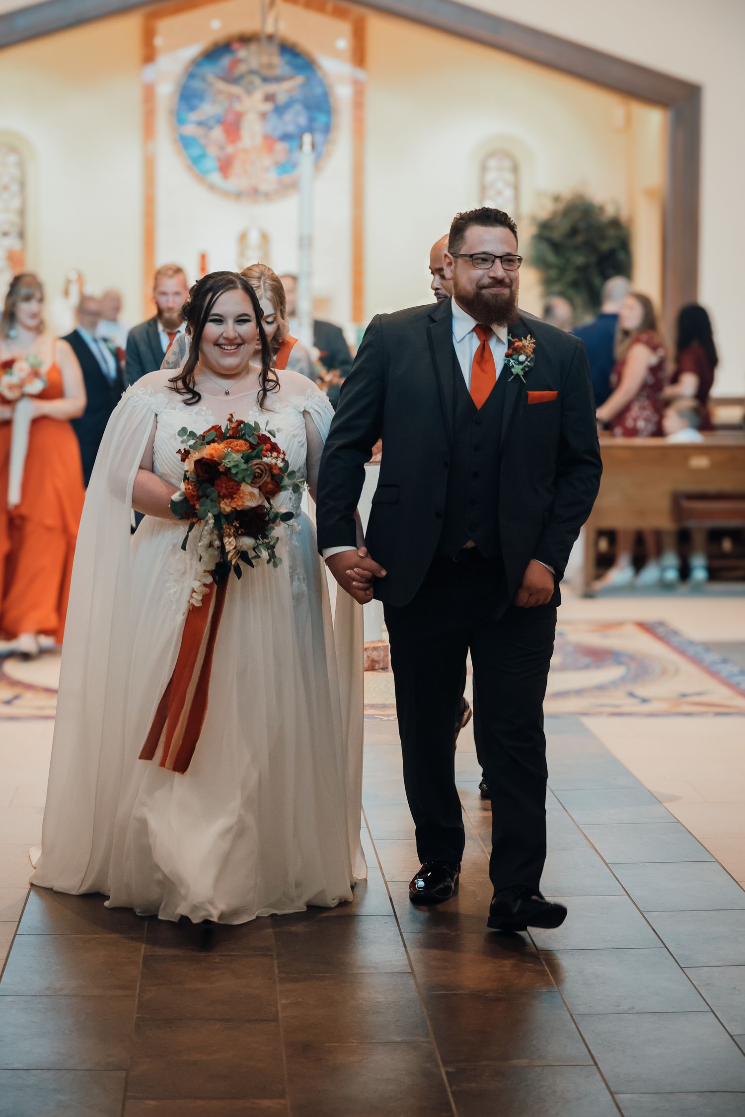 Wedding party members in black suits and cream dresses walk down a church aisle with autumn-colored floral bouquets.