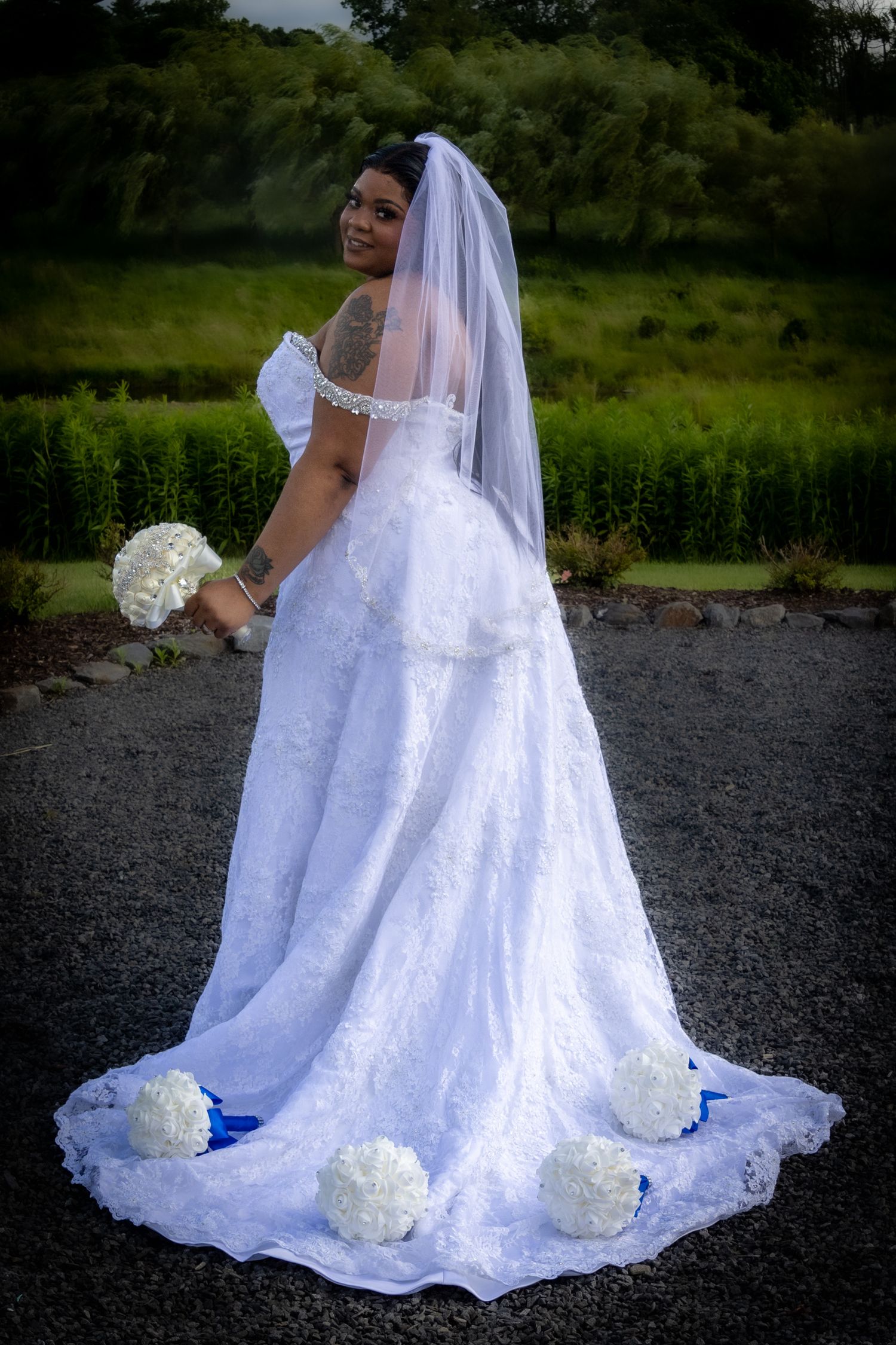 A bride in a flowing white lace wedding gown with a veil sits on dark ground while holding a white bouquet.