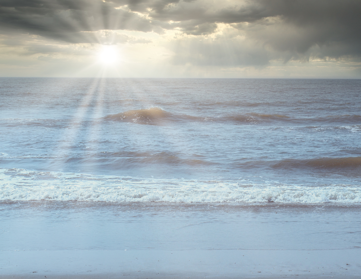Sunbeams breaking through dramatic storm clouds over calm ocean waves on a sandy beach.