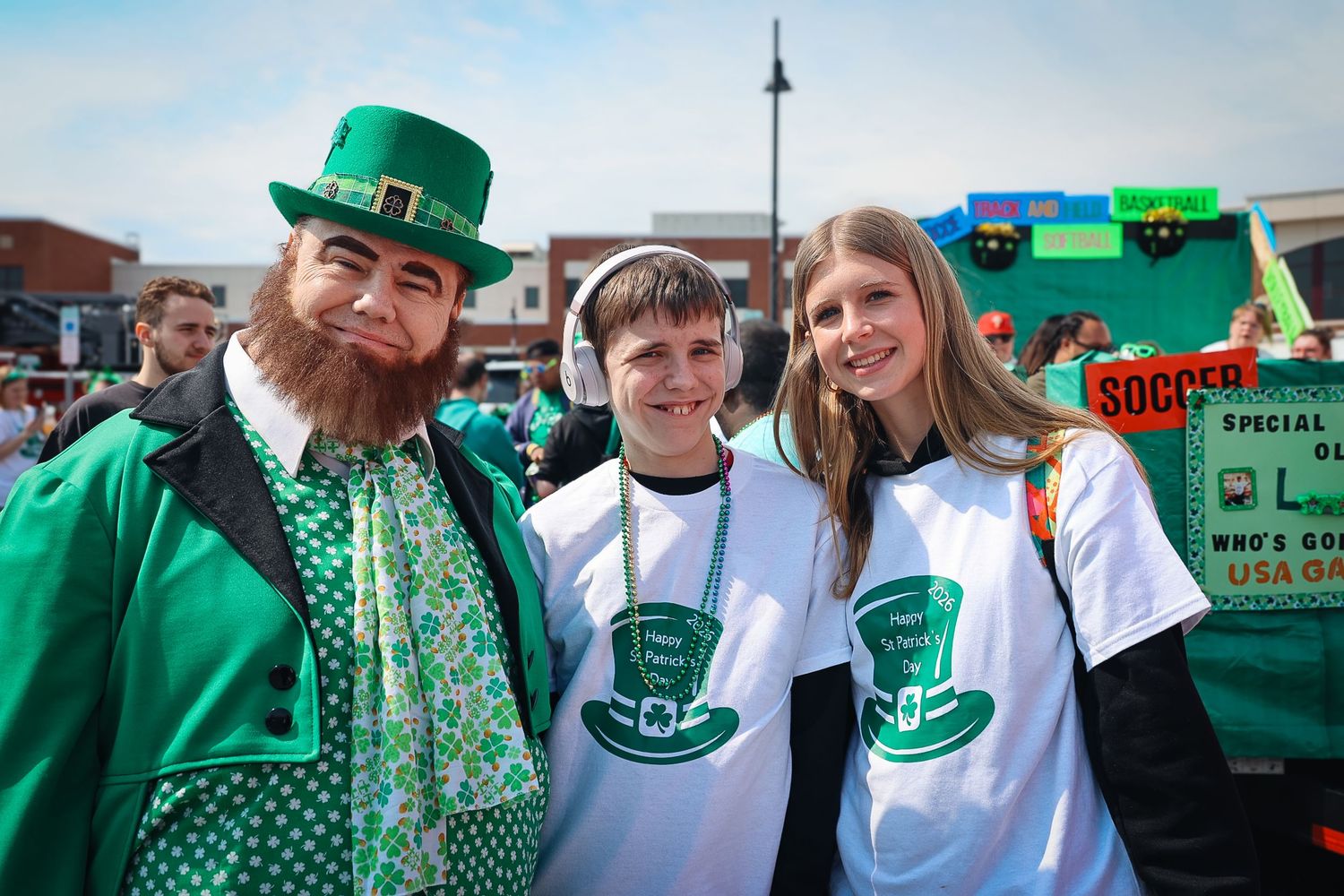Three people in green St. Patrick's Day outfits pose together outdoors, one dressed as a leprechaun with a red beard.