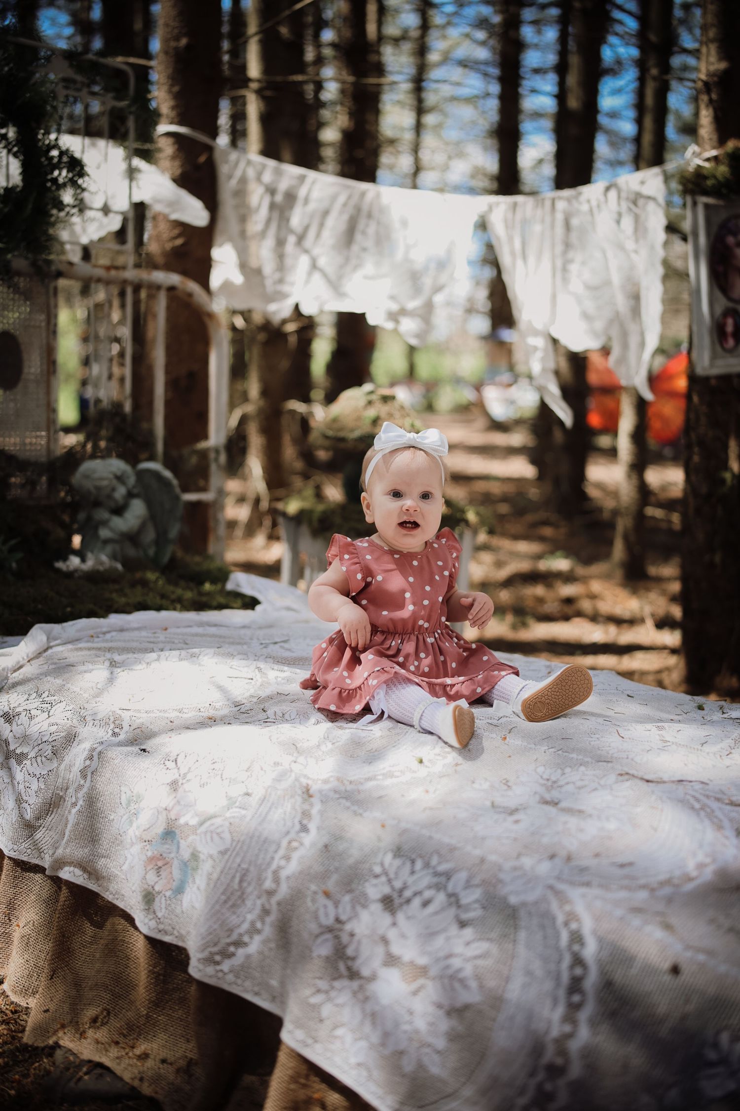 Baby girl in pink dress sits on white lace blanket outdoors in forest with laundry line behind her.