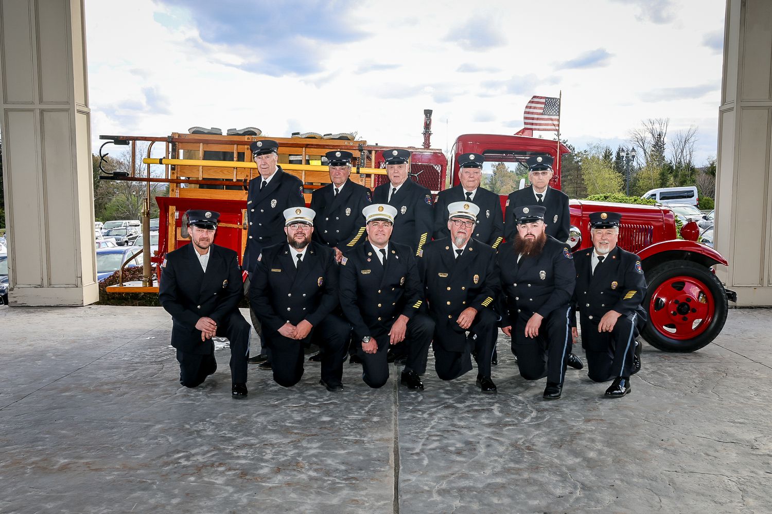 Firefighters in dress uniforms pose in front of a vintage red fire truck at a fire station.