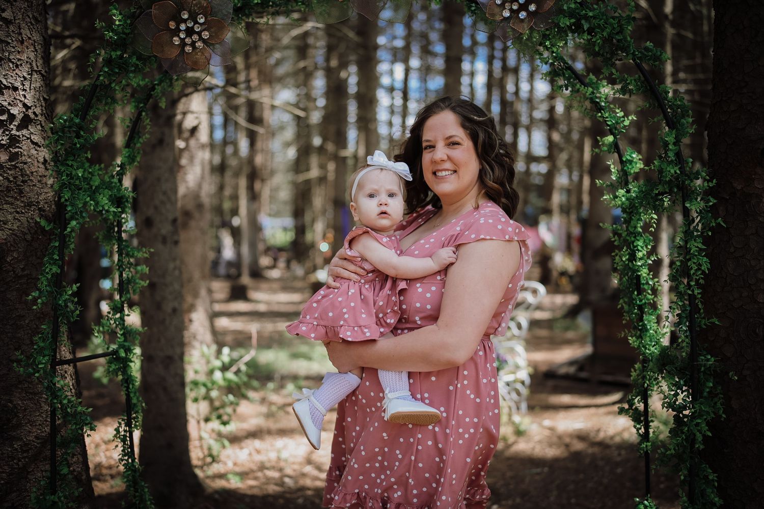 A woman in a pink floral dress holds a baby girl in matching outfit under a floral arch in a forest setting.