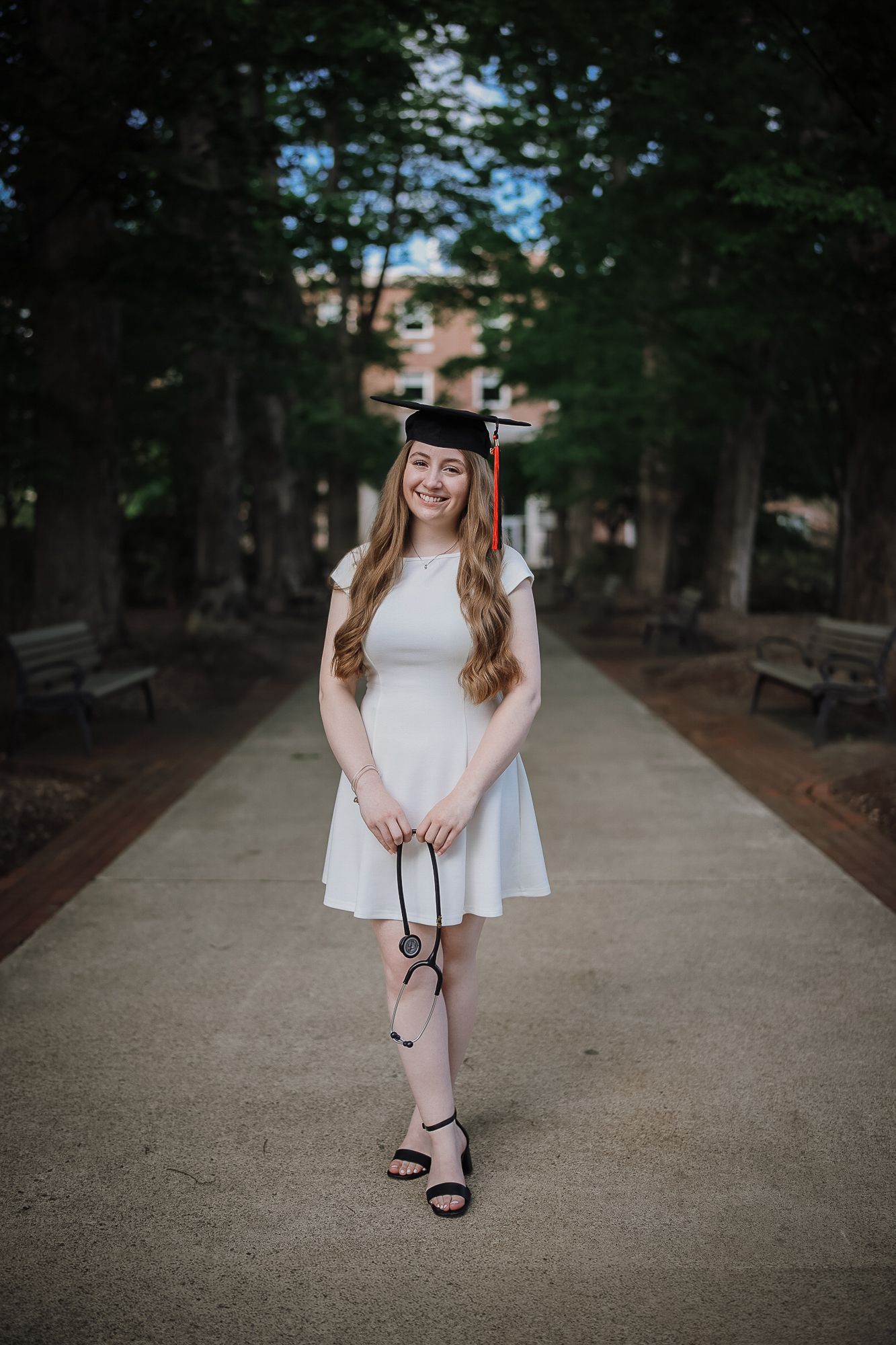 Young woman in white dress and graduation cap smiling on a tree-lined campus walkway, holding sunglasses.