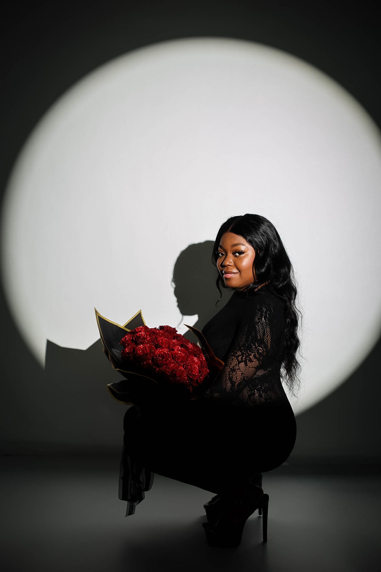 Woman in black lace outfit kneeling with bouquet of red roses against dramatic circular spotlight background.