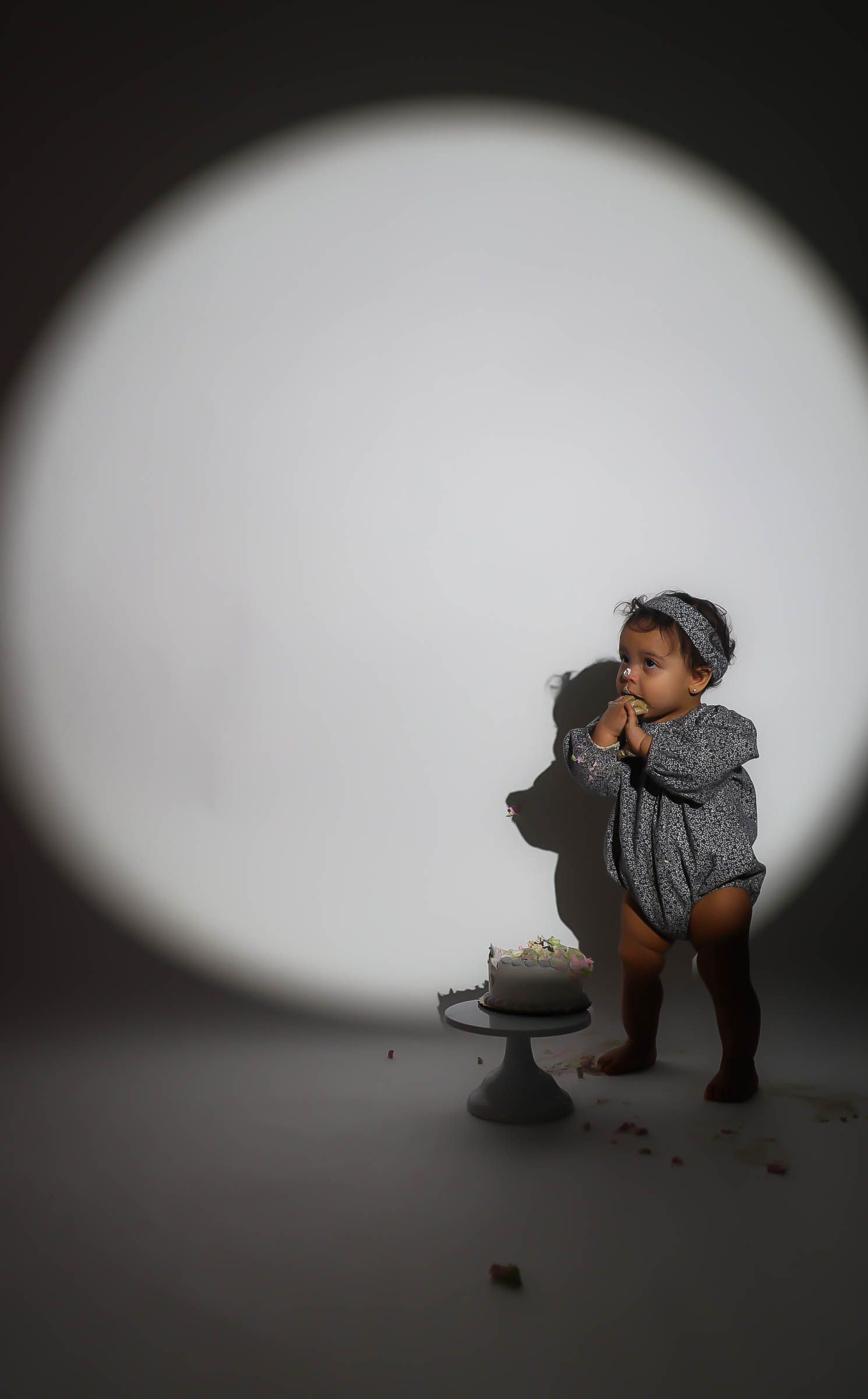 A toddler in a gray romper stands by a smashed birthday cake under a circular spotlight.