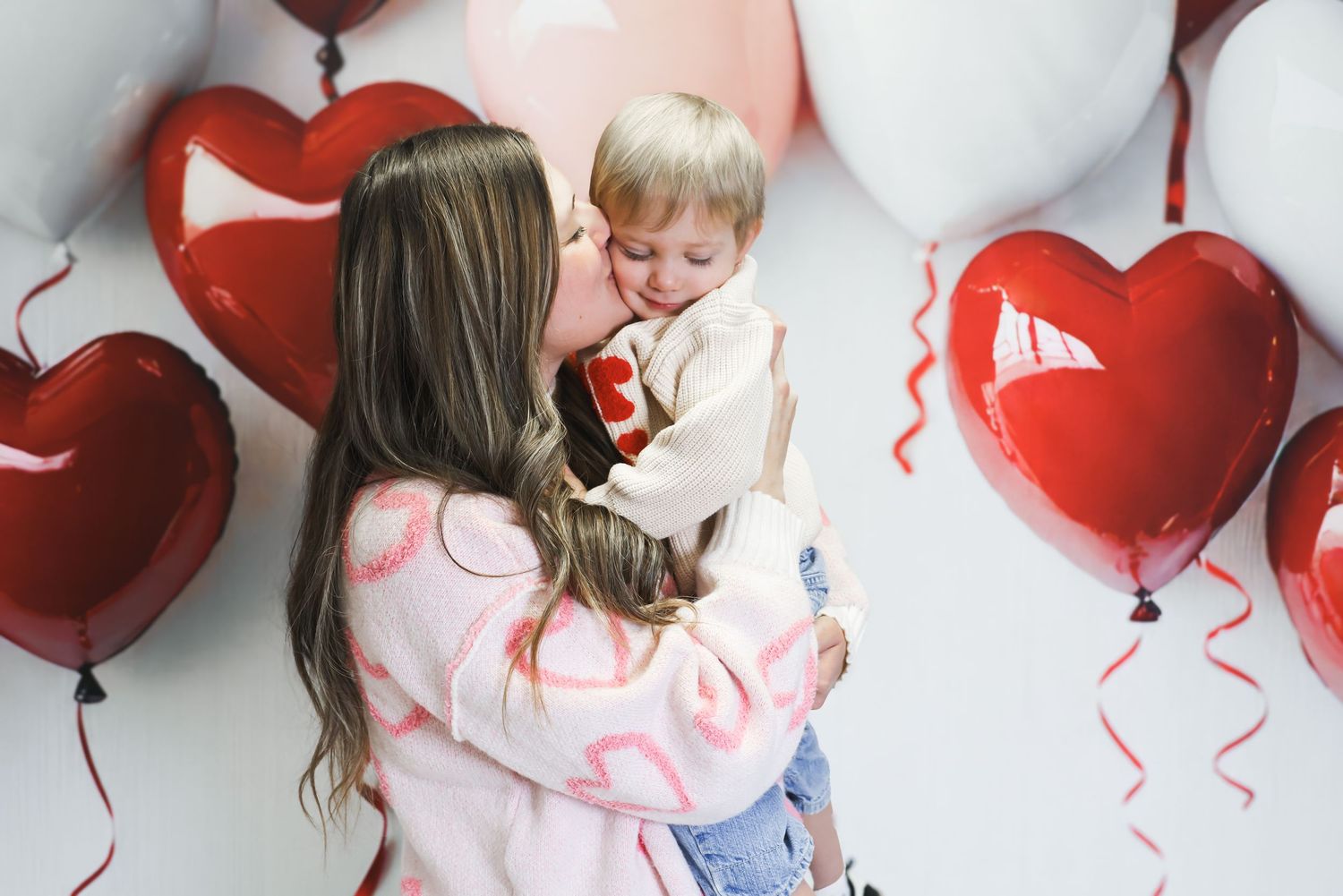 A mother kisses her toddler surrounded by red heart-shaped balloons on a white background.